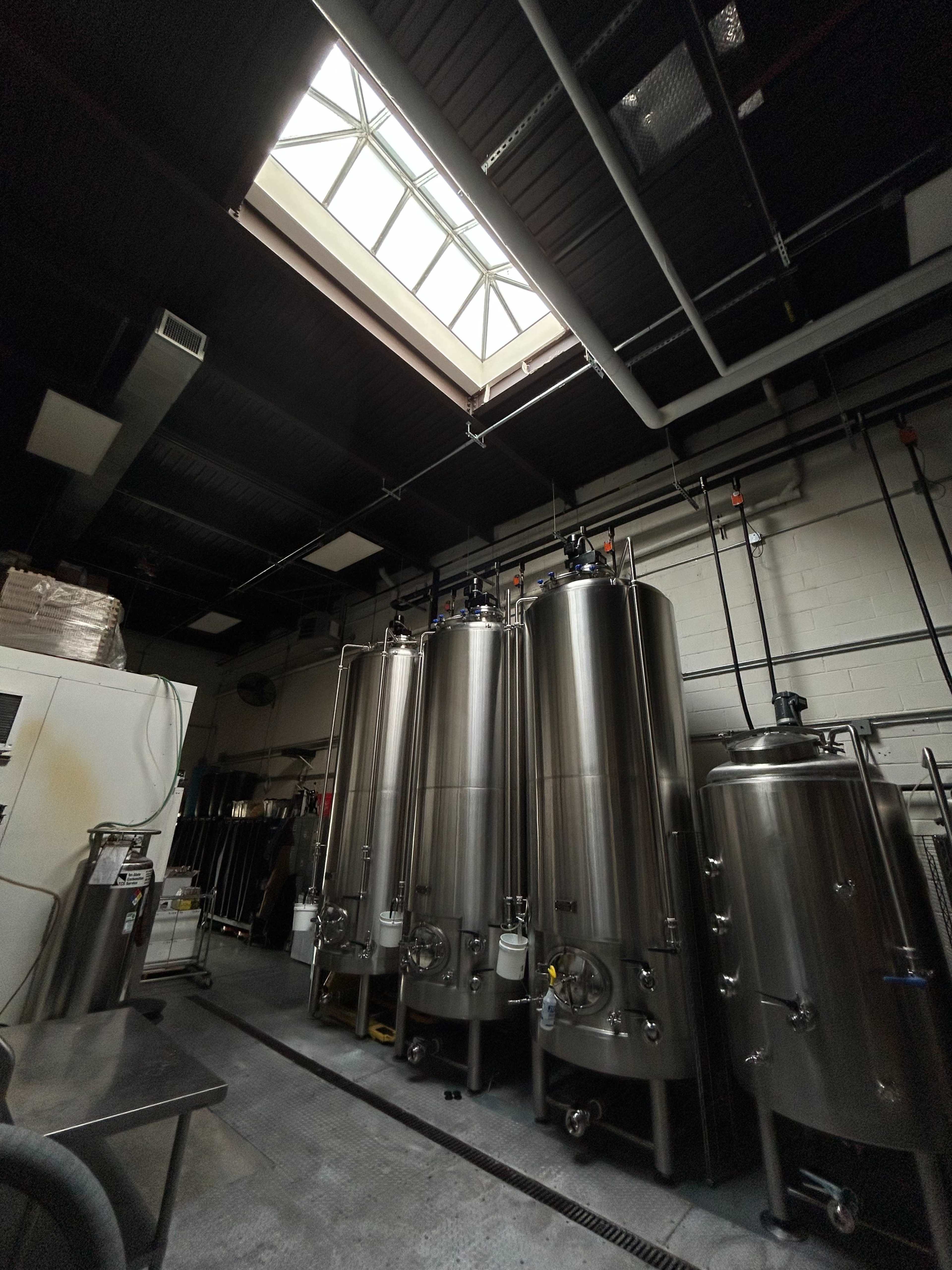 The image shows a room with several large stainless steel fermentation tanks and a skylight above, providing natural light.