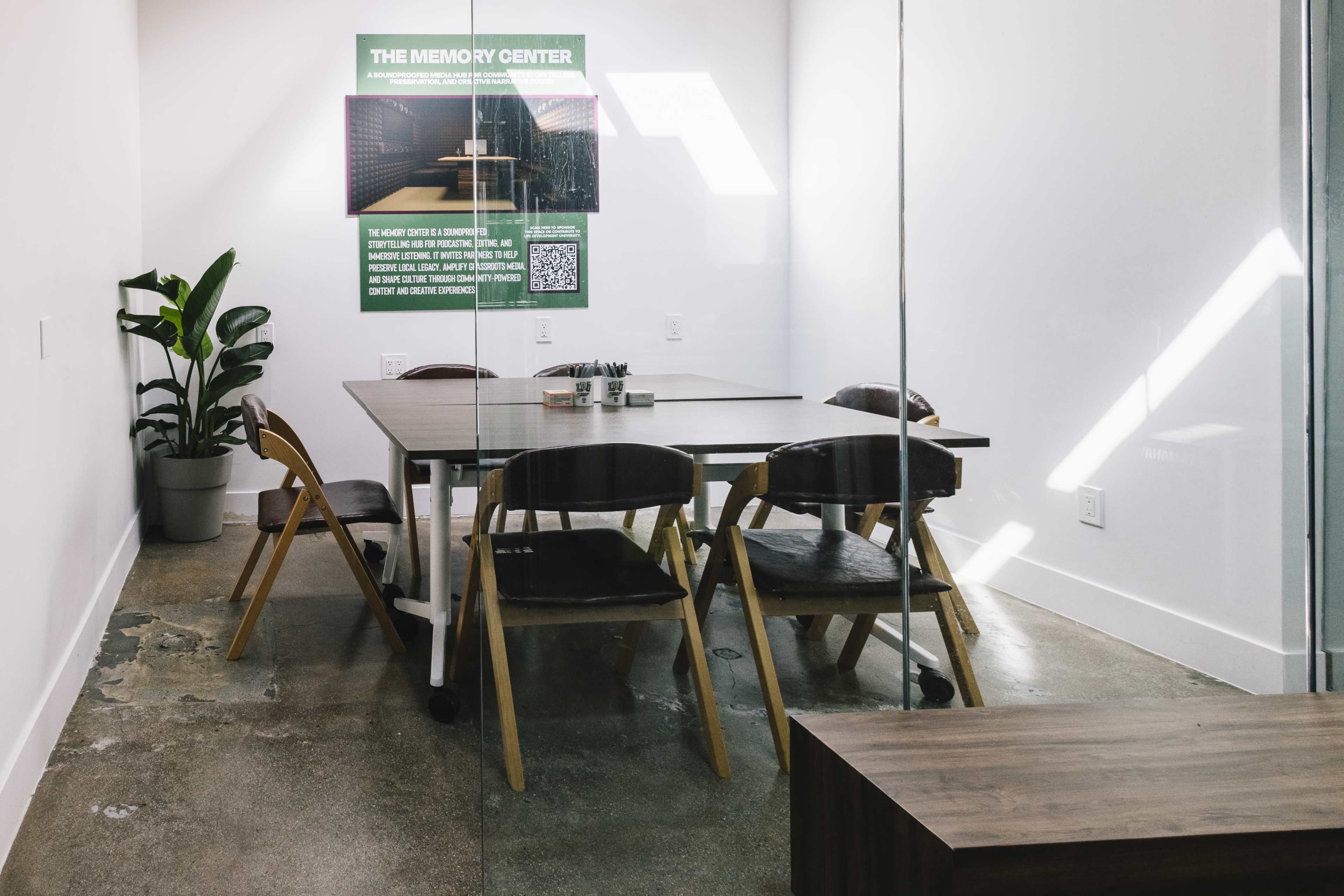 A modern conference room features a large table surrounded by six wooden chairs, with a plant in the corner and a poster on the wall.