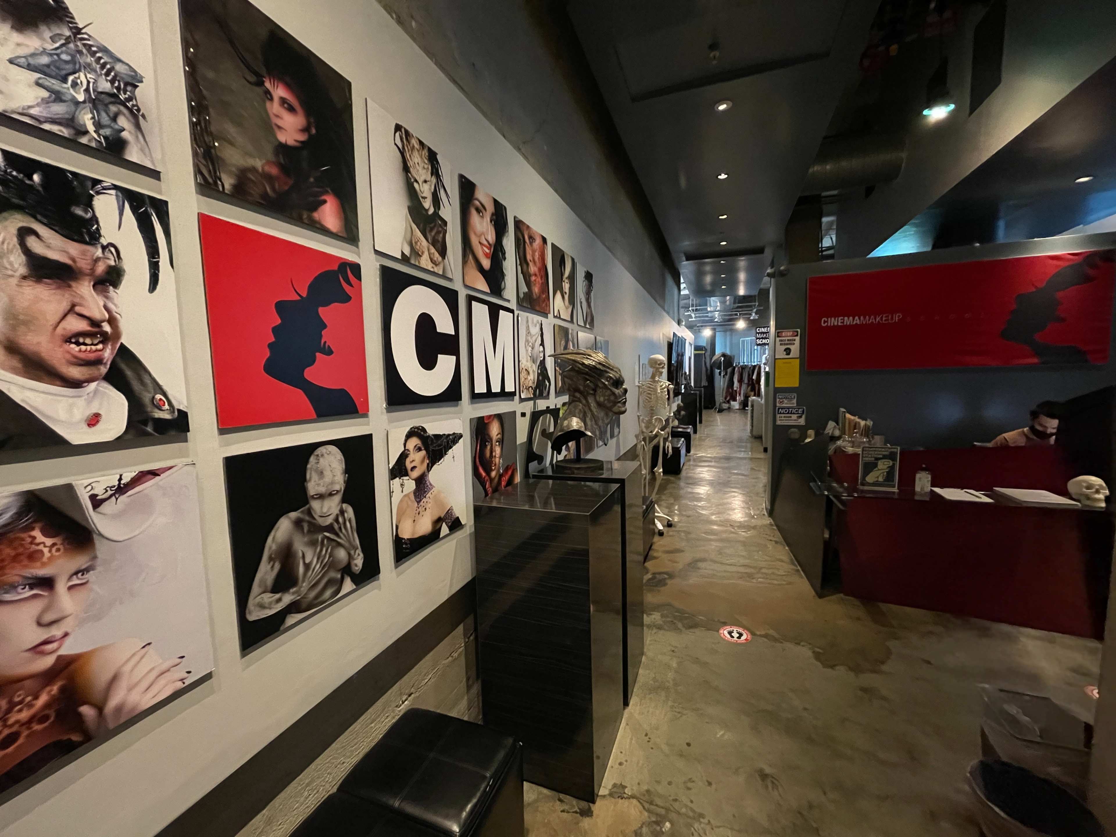 The image shows a narrow hallway adorned with a series of framed artistic portraits on one side and a reception area with a desk on the other.