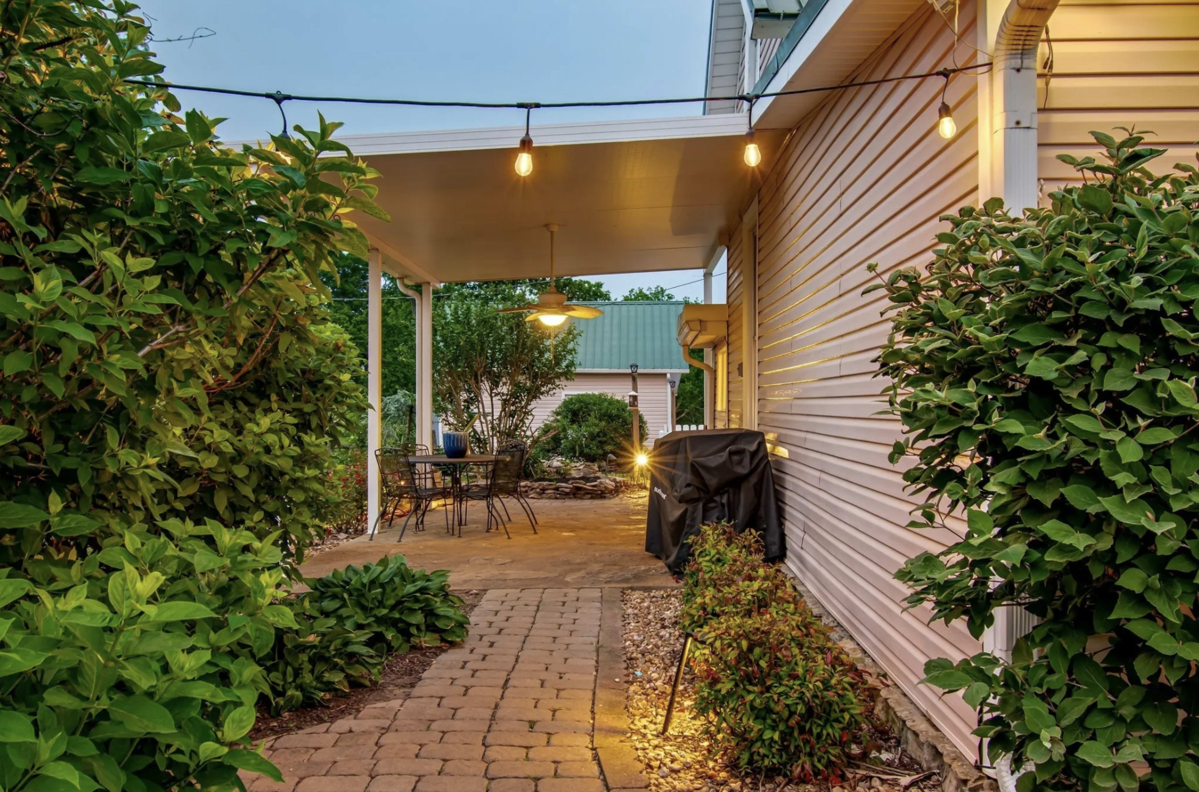 A stone pathway leads through landscaped greenery to a covered patio area with outdoor seating and hanging lights.