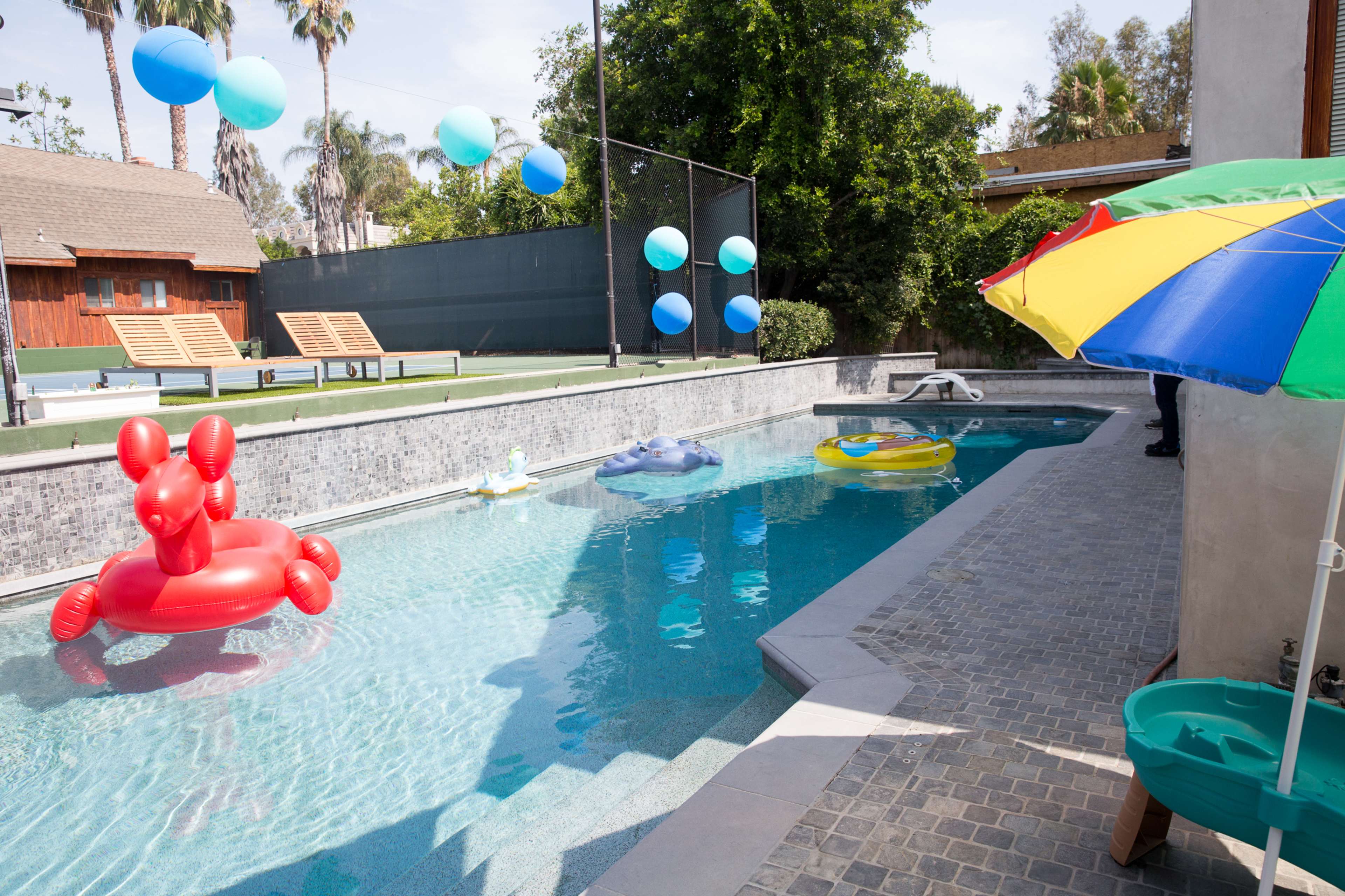 A swimming pool features inflatable floats and colorful balloons, with lounge chairs beside the water and a shaded umbrella nearby.