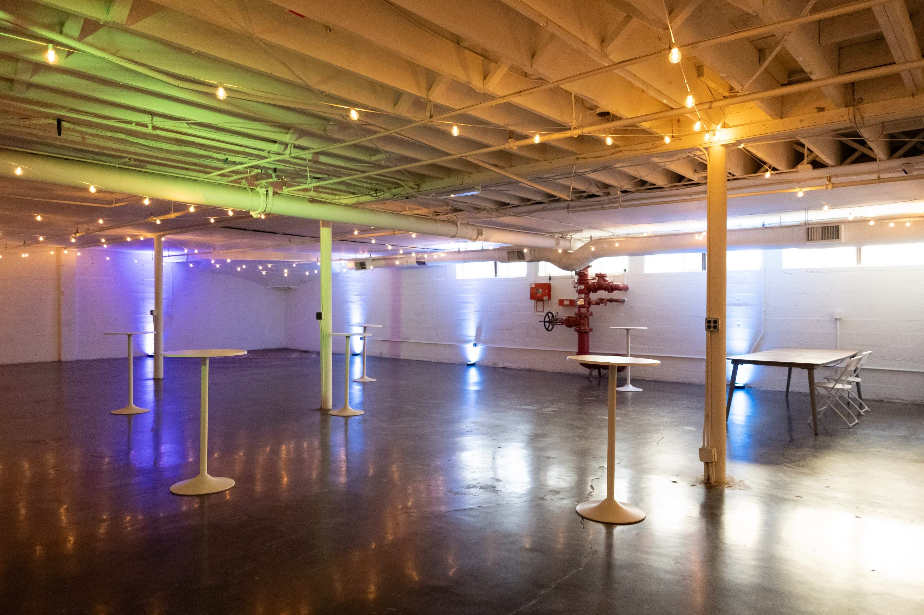 The image shows a spacious, well-lit event venue with white walls, polished concrete floors, and several tall tables, enhanced by string lights and purple lighting effects.