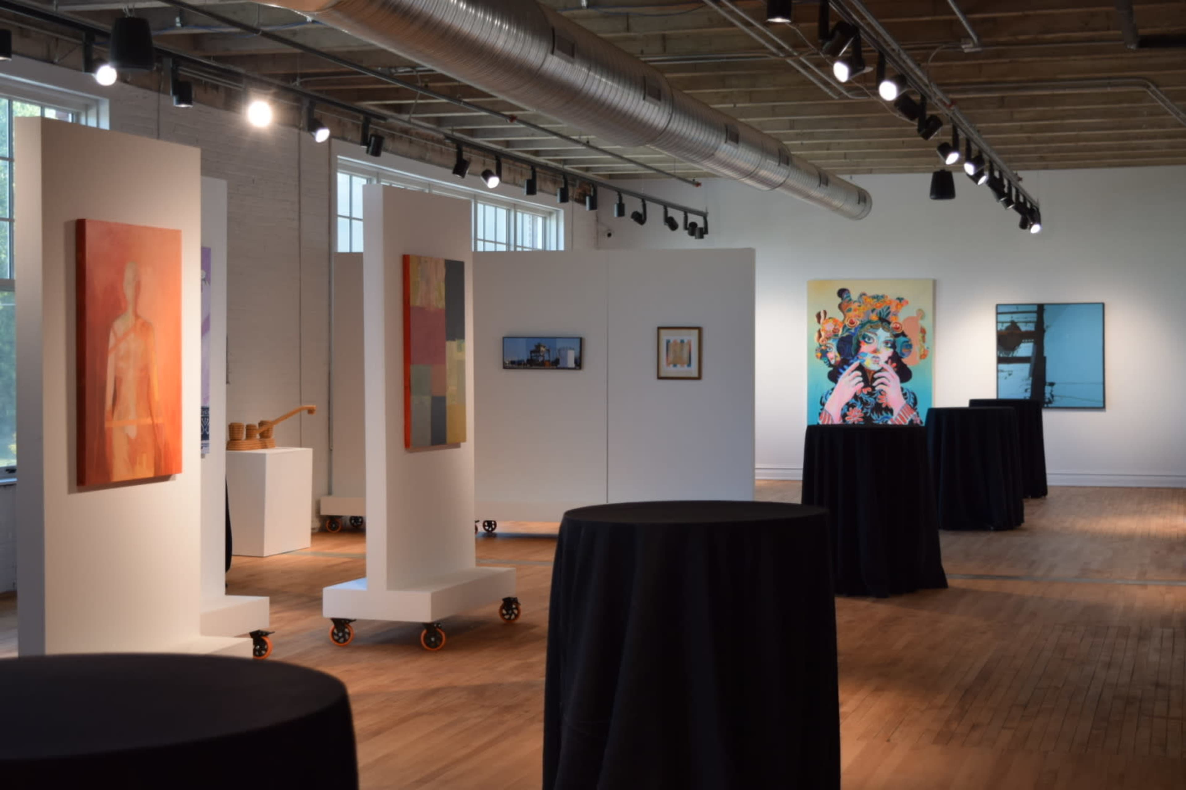 The image shows an art gallery interior with white panels displaying various artworks and black tables placed around the space.