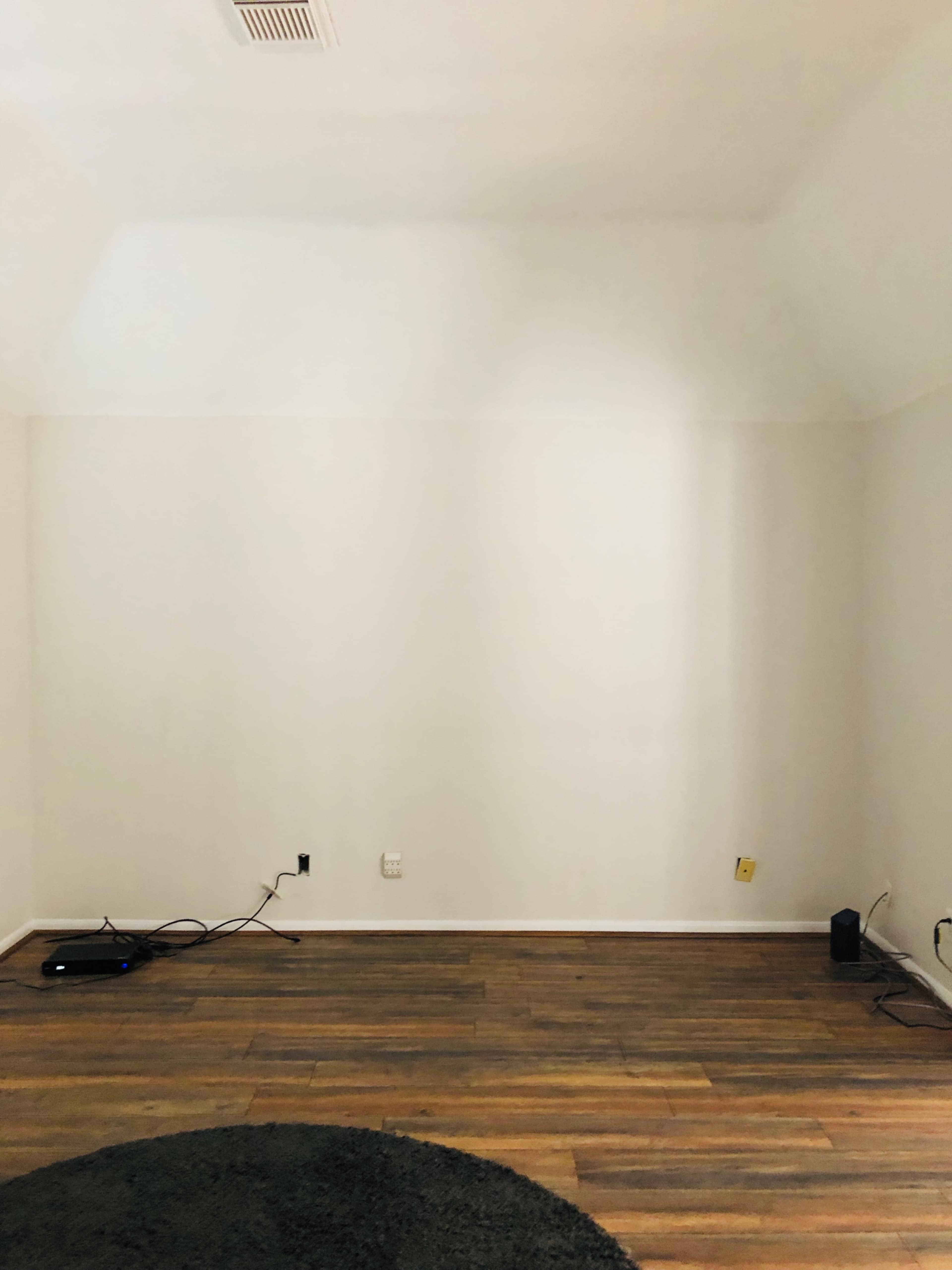 An empty room with light-colored walls and wooden flooring, featuring a circular rug on the floor and some cables along the wall.