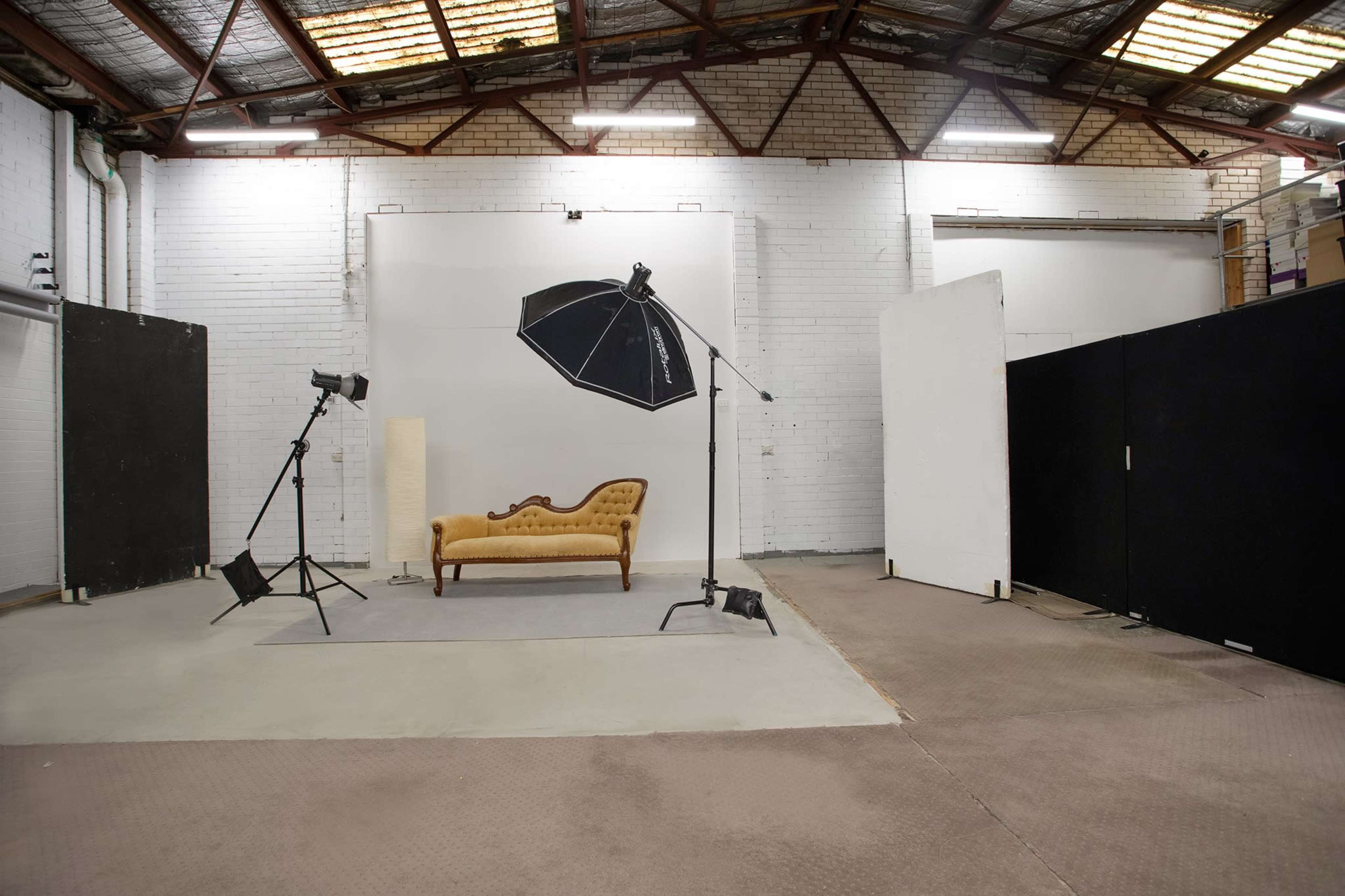A photo studio featuring a vintage-style couch, a large umbrella light, and various backdrops in a spacious, industrial setting.