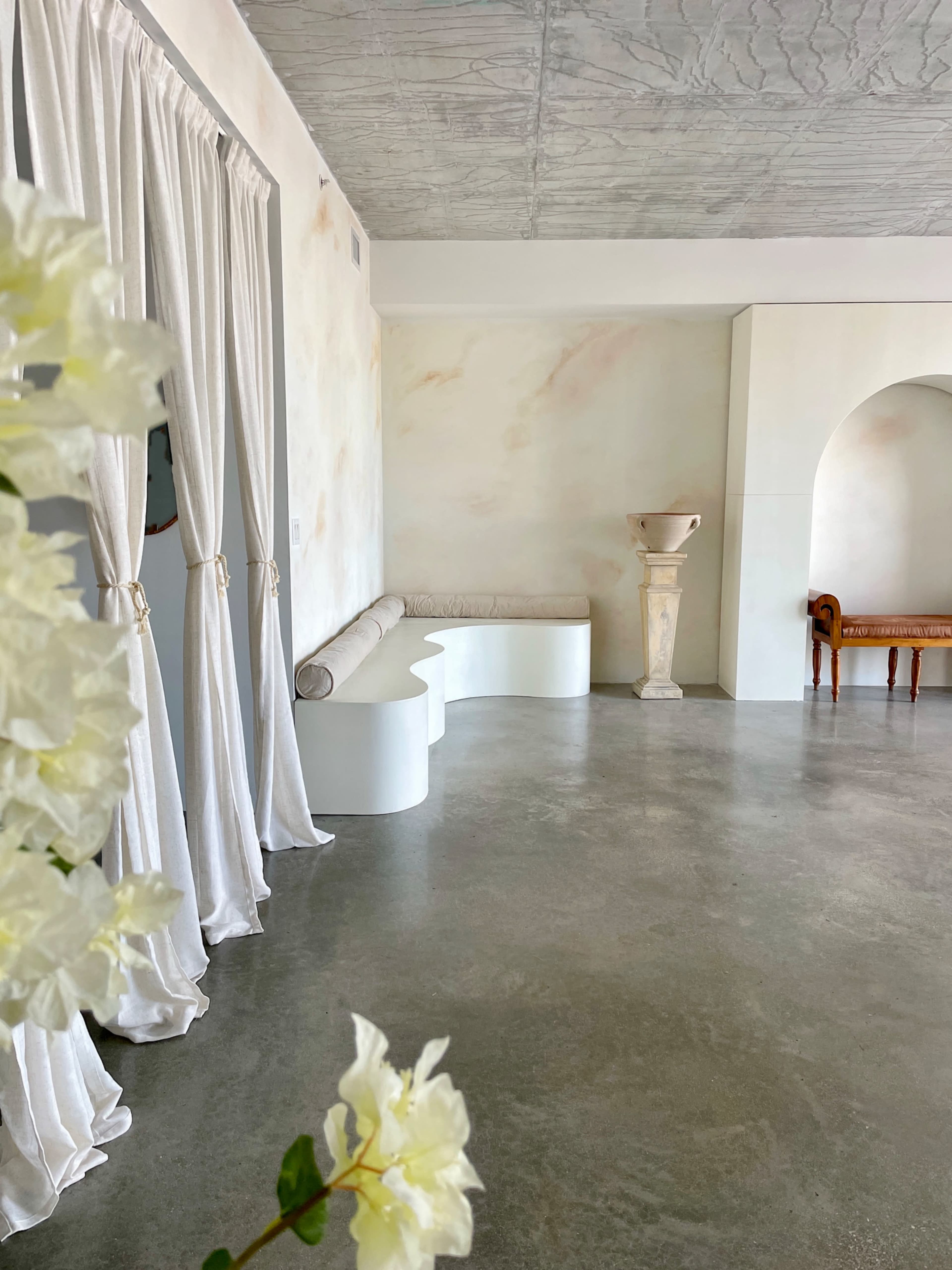 The image shows a minimalist interior space with a curved white bench, a decorative column, and a wooden chair beside sheer curtains.