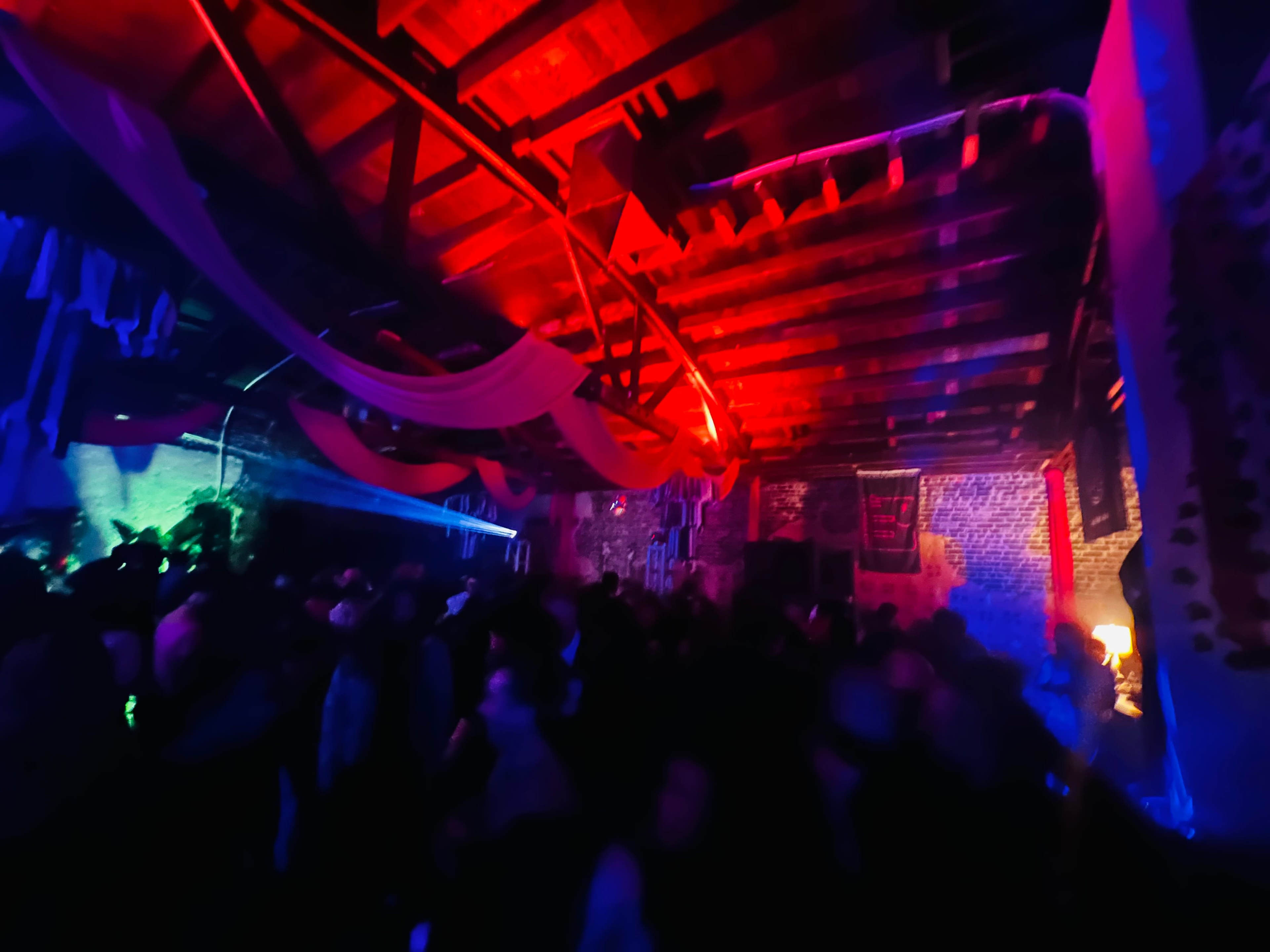 A dimly lit party is happening inside a warehouse, with colorful lights illuminating the crowd and decorations hanging from the ceiling.