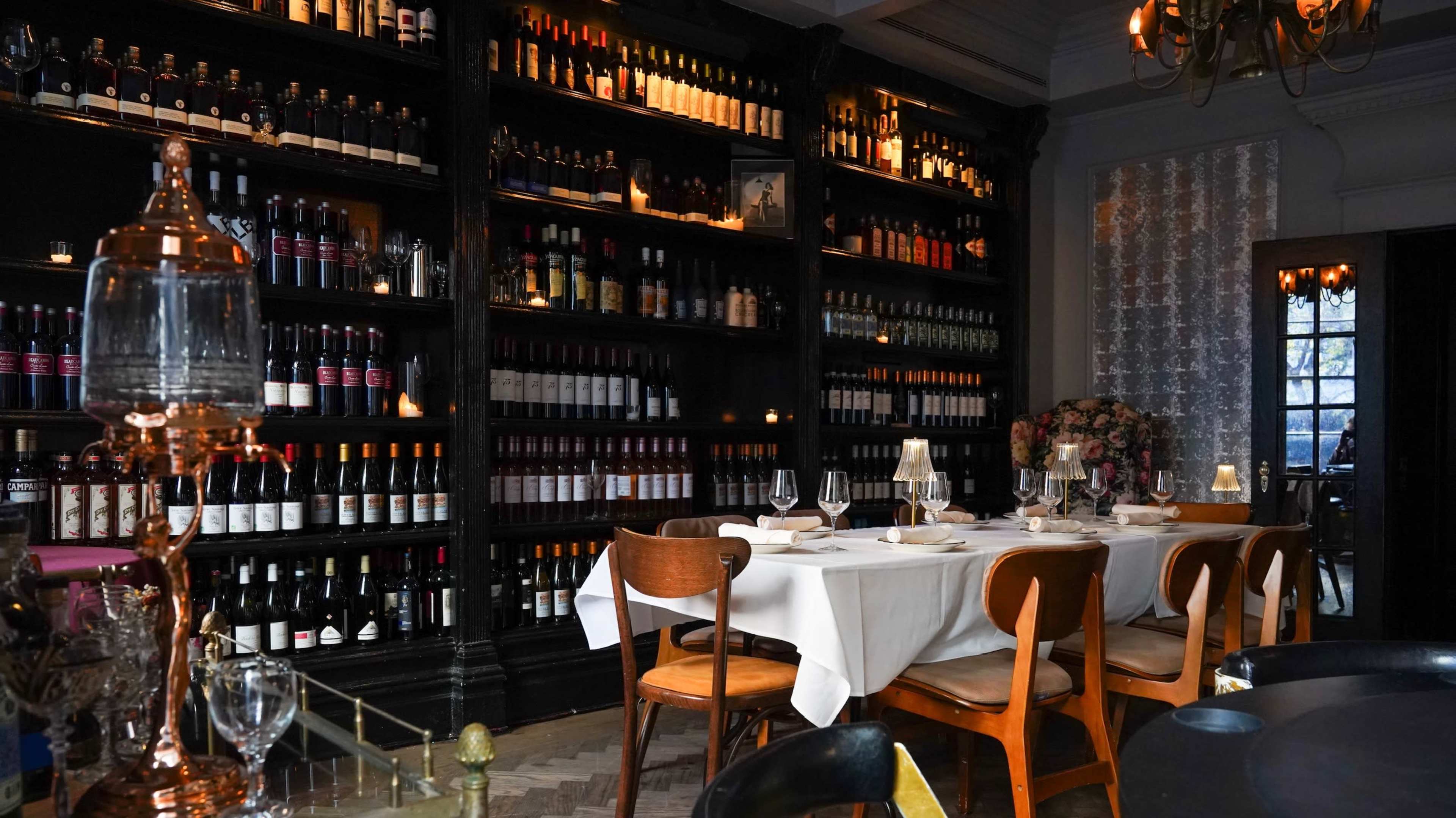 A table is set for dining in a dimly lit restaurant, with a large bookshelf filled with bottles of wine in the background.