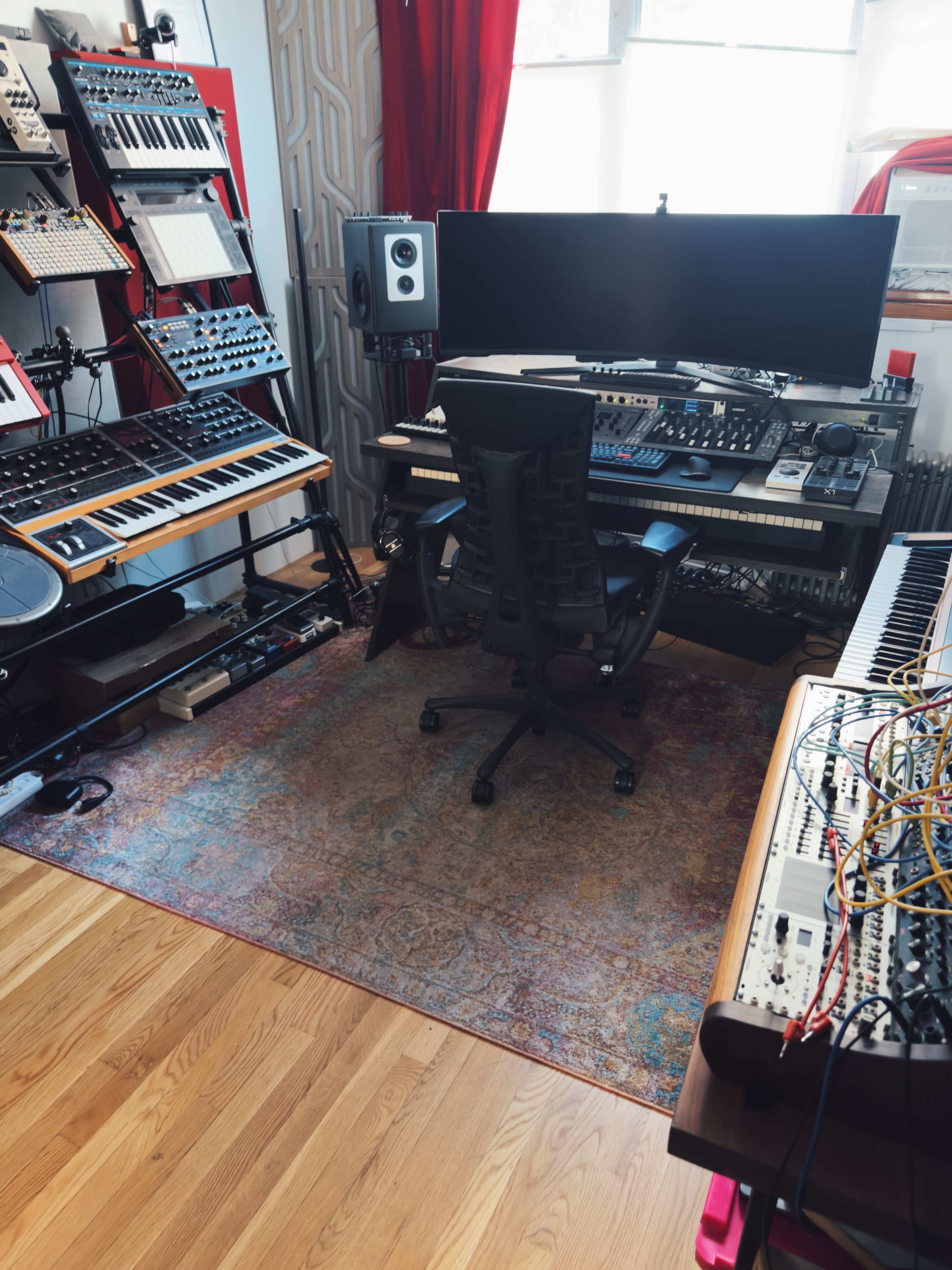 The image shows a music production studio with various synthesizers, a computer monitor, and a central desk chair on a patterned rug.