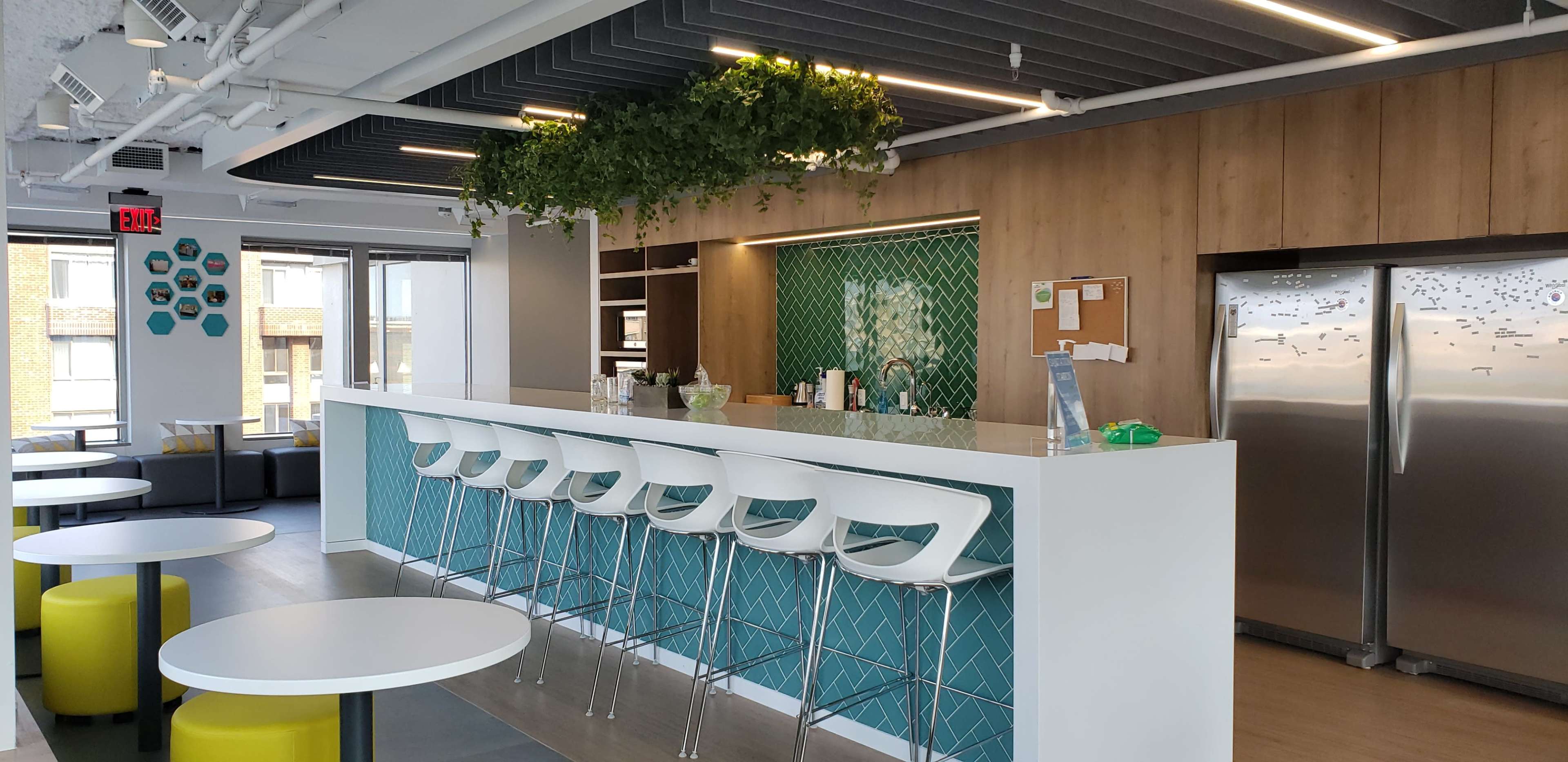 A modern kitchen area with a bar counter, high stools, and green wall decor, along with a refrigerator and a seating area in the background.