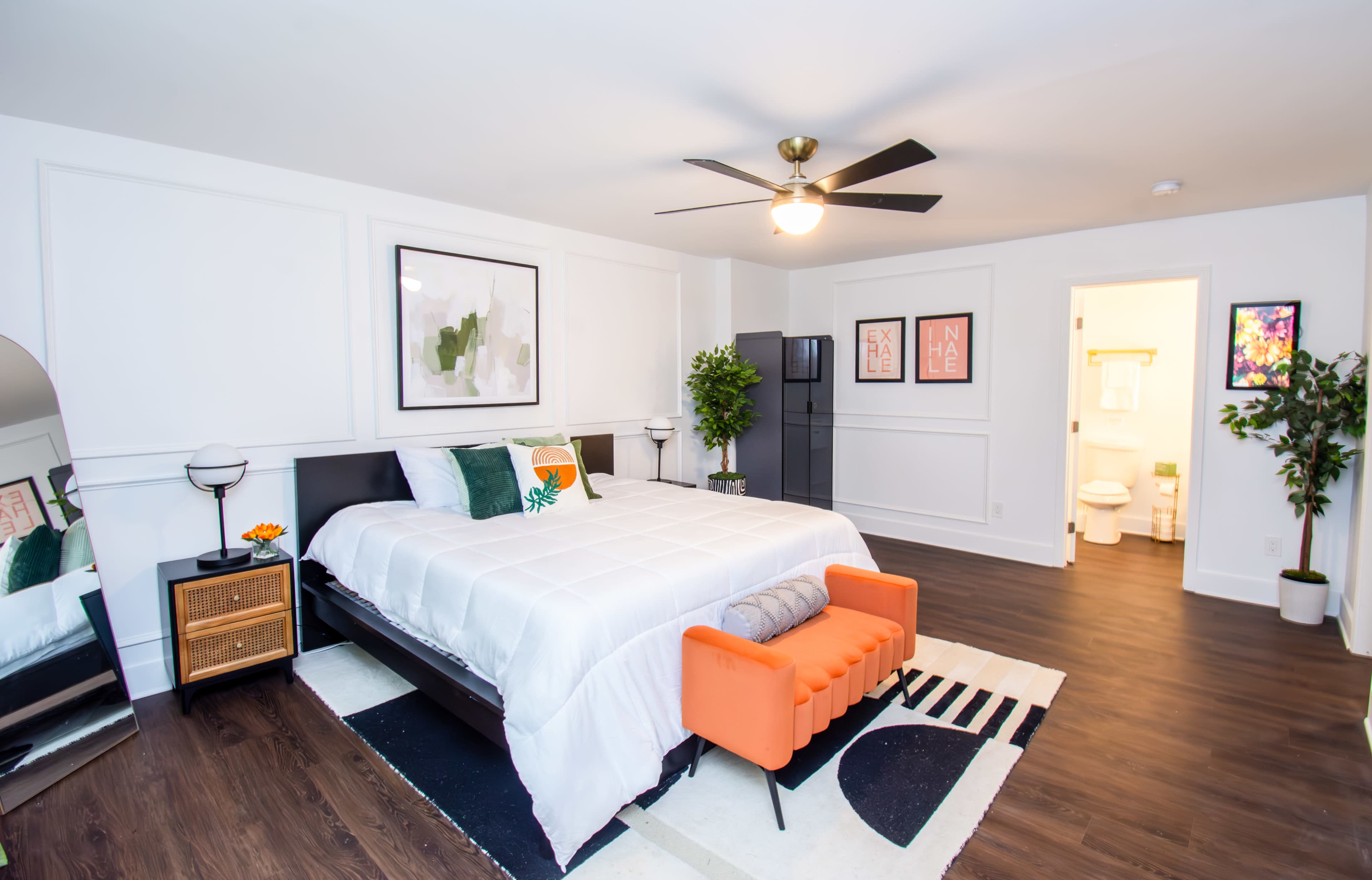 The image shows a modern bedroom with a large bed, a bright orange accent chair, and a bathroom visible through an open door.