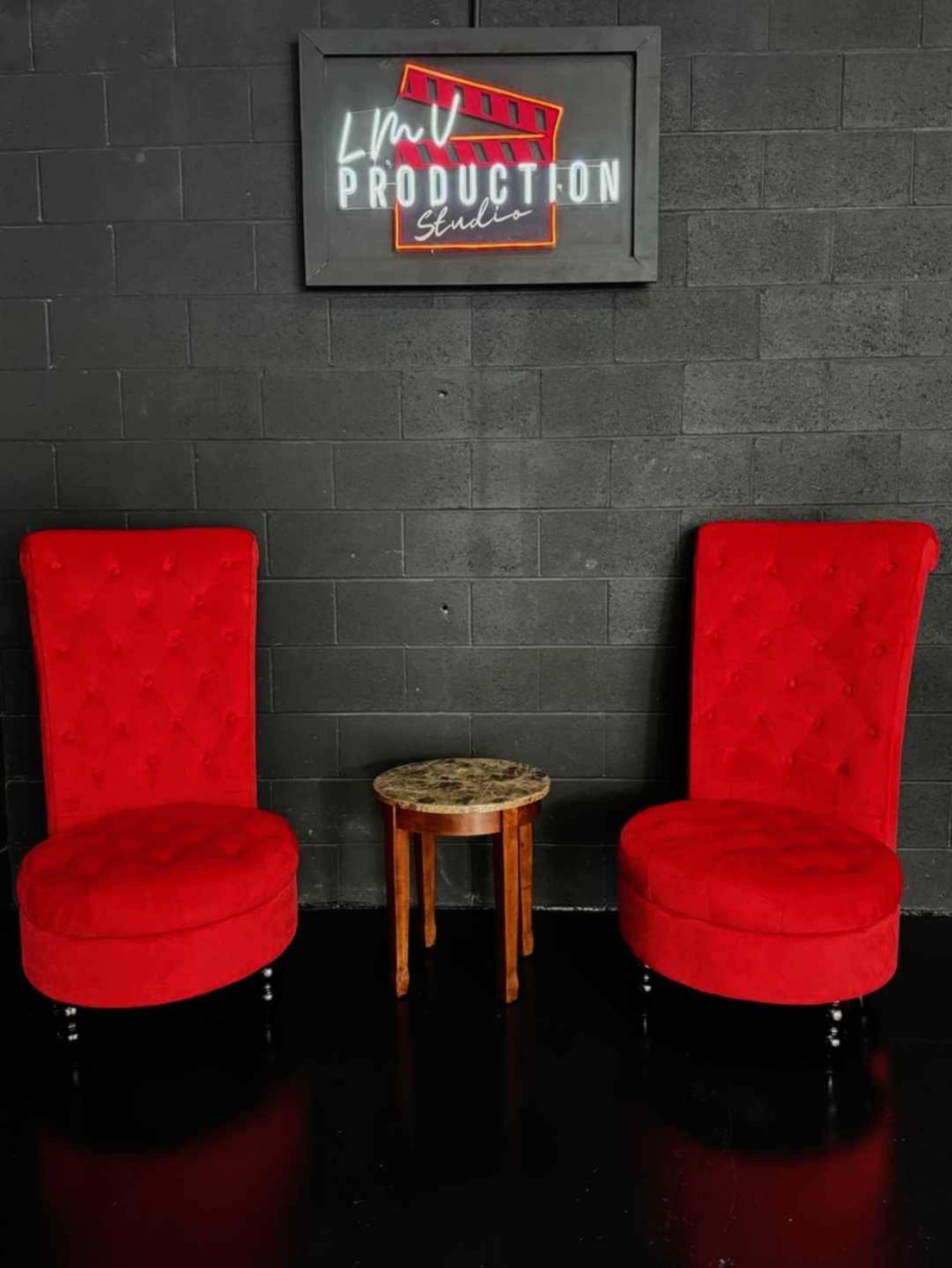 Two red velvet chairs and a small wooden table set against a black wall with a sign that reads "LMV PRODUCTION Studio."