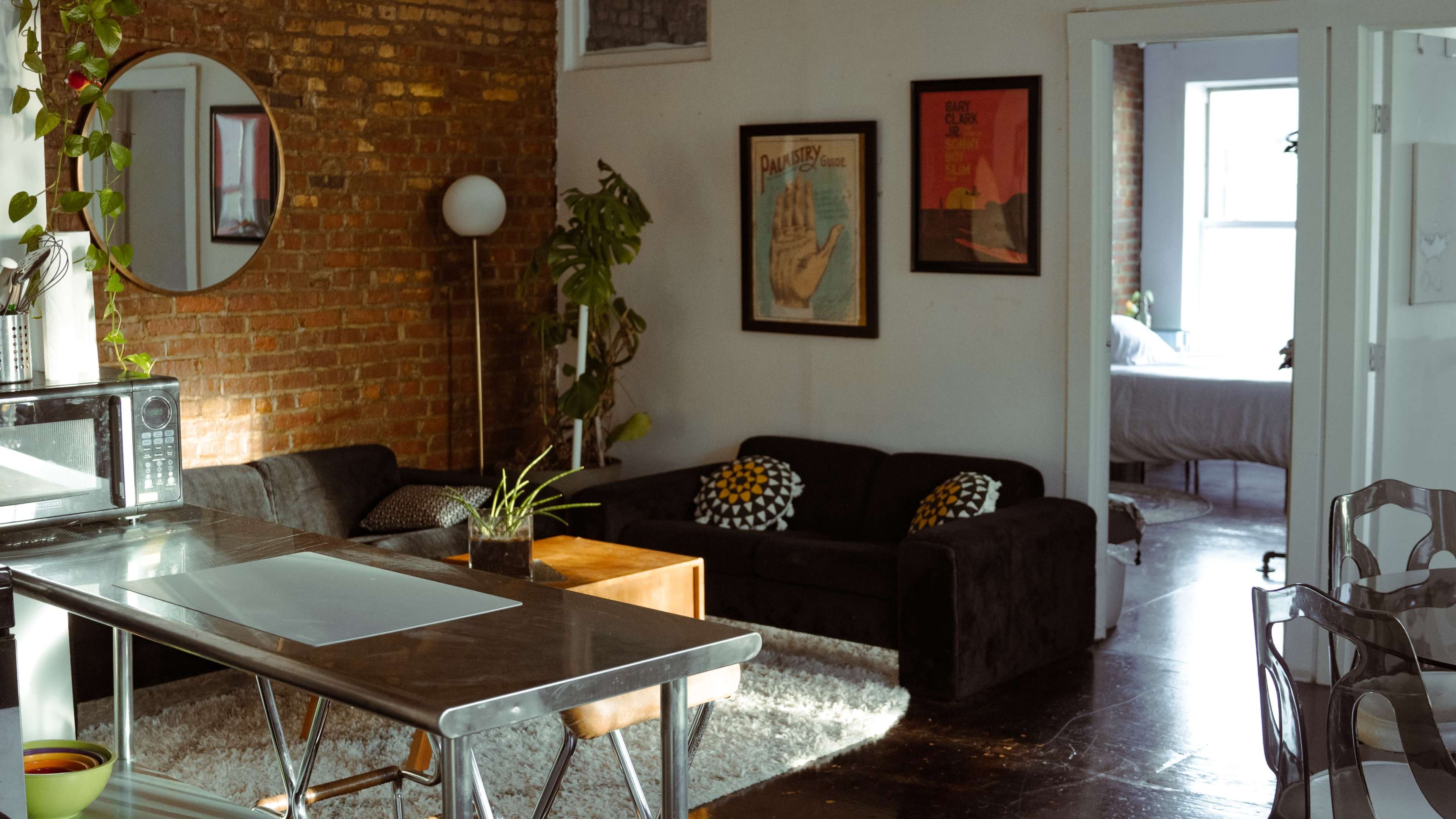 A modern living space features a kitchen area with a stainless steel table and microwave, adjacent to a cozy seating area with dark sofas and framed artwork on the exposed brick wall.