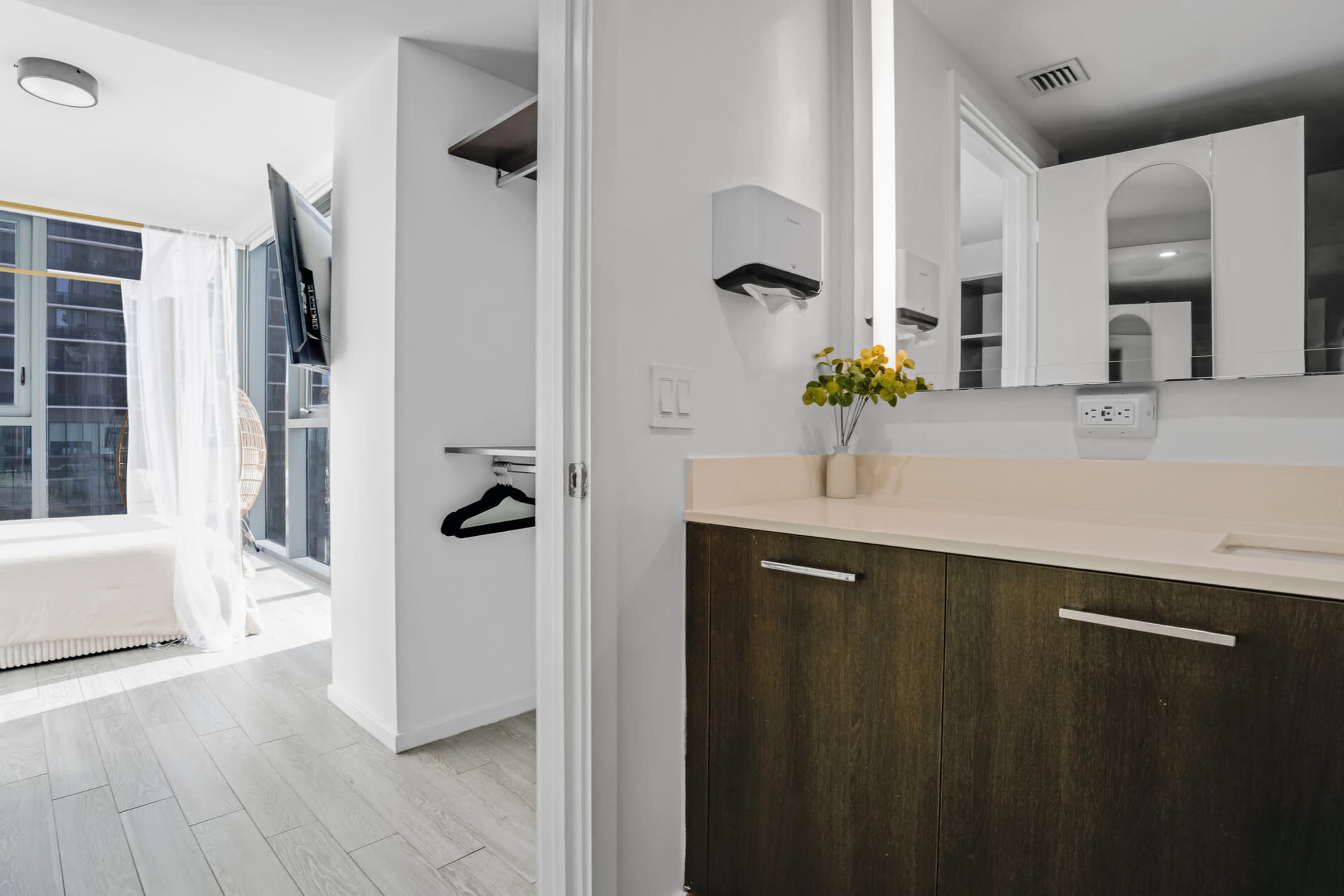 The image shows a modern bathroom area with a sink and wooden cabinetry, leading into a light-filled space featuring a bed and a television.