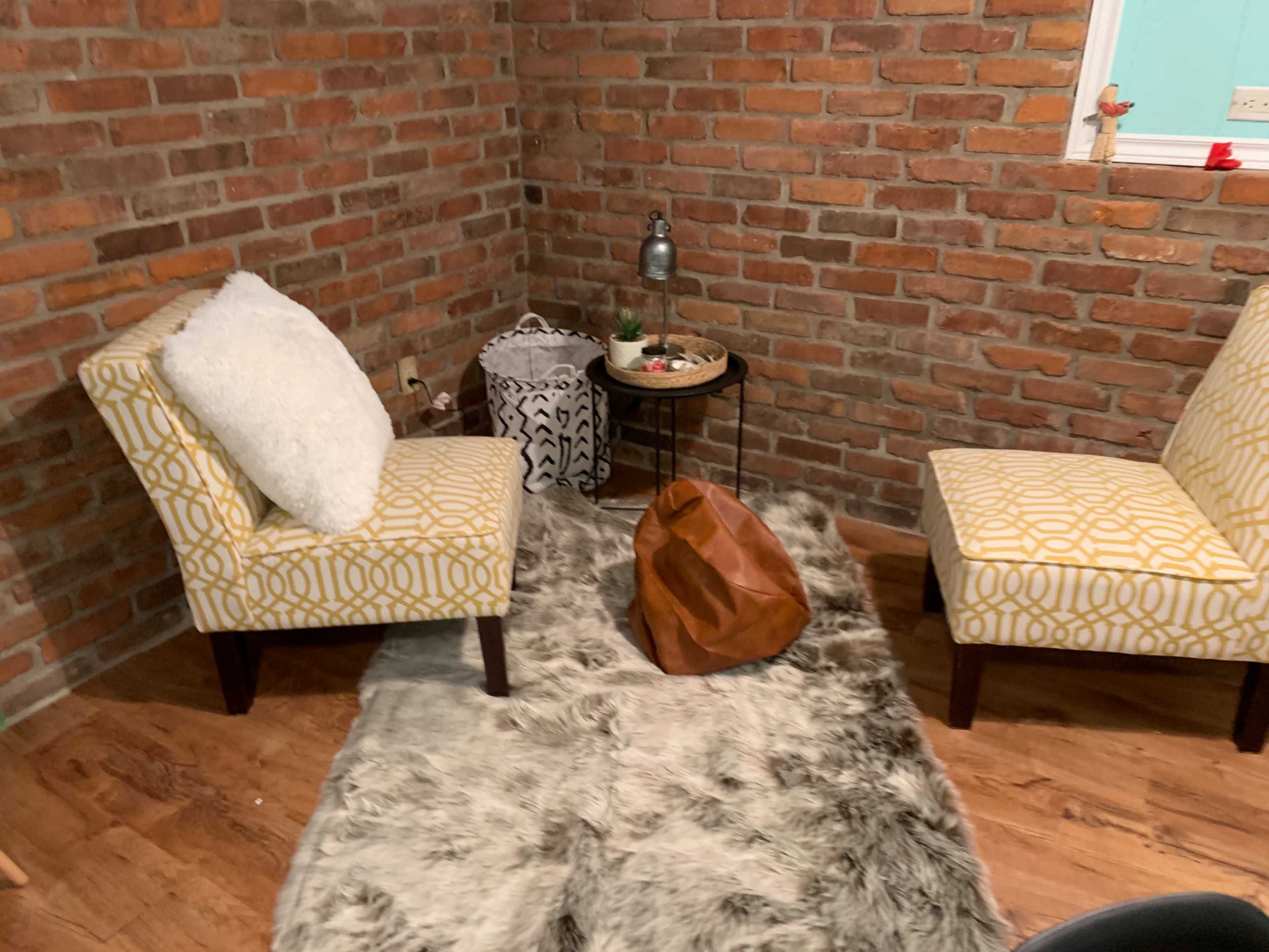 A cozy corner with two patterned chairs, a round side table, a decorative bowl, and a brown bean bag on a faux fur rug against a brick wall.
