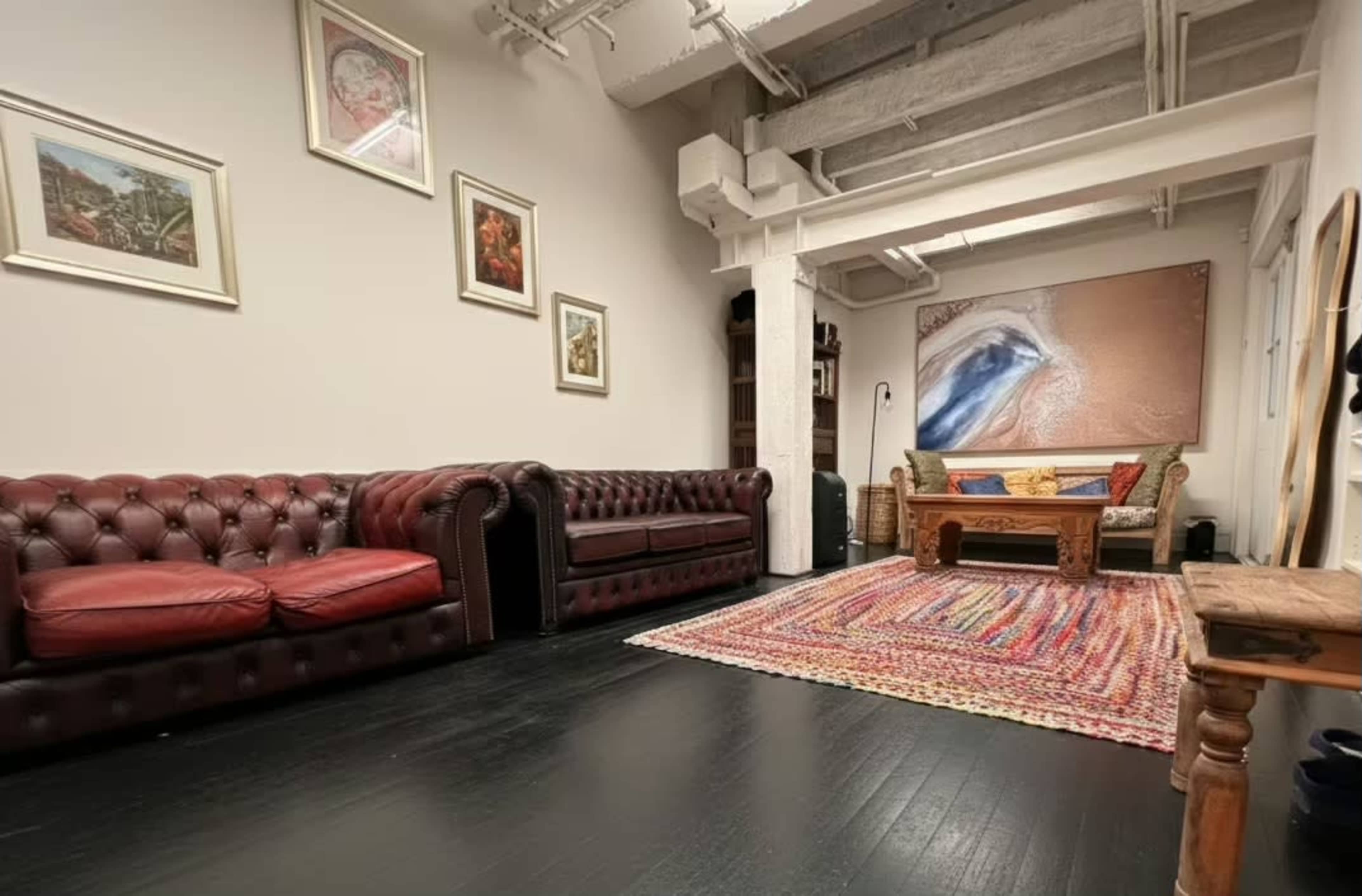 The image shows a spacious room with two red leather chesterfield sofas, a wooden coffee table, a colorful area rug, and artwork on the walls.
