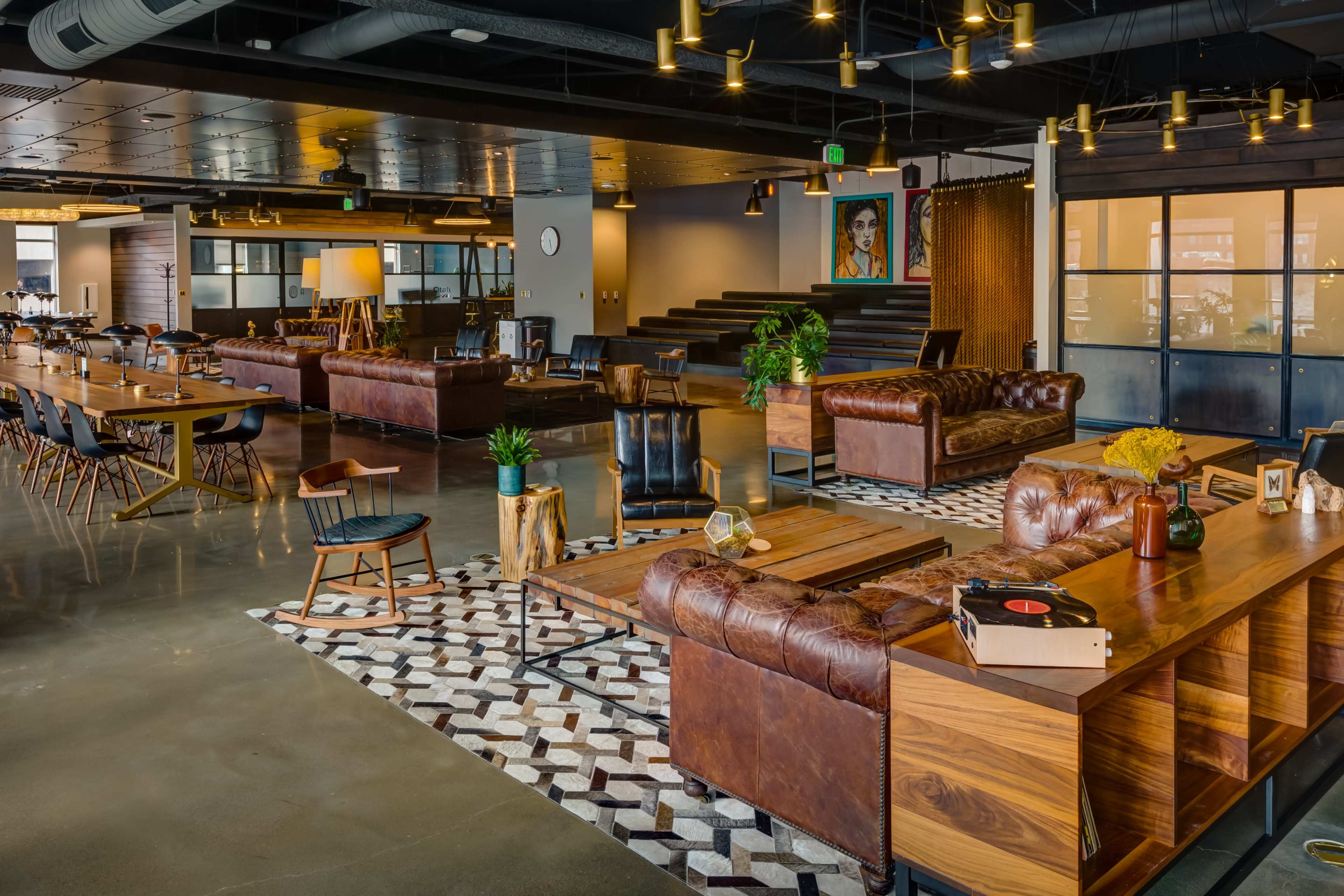 A modern office lounge area with leather sofas, wooden tables, and a patterned rug, featuring a large mural on the wall and various plants.