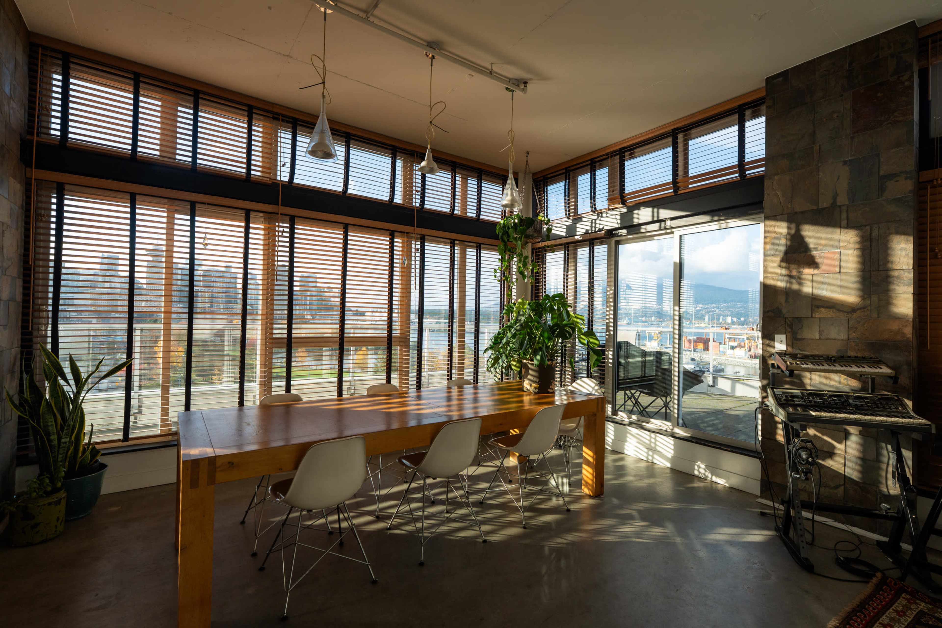 The image shows a modern, well-lit interior space featuring a wooden dining table surrounded by chairs, large windows with blinds, and plants, with a view of a waterfront outside.
