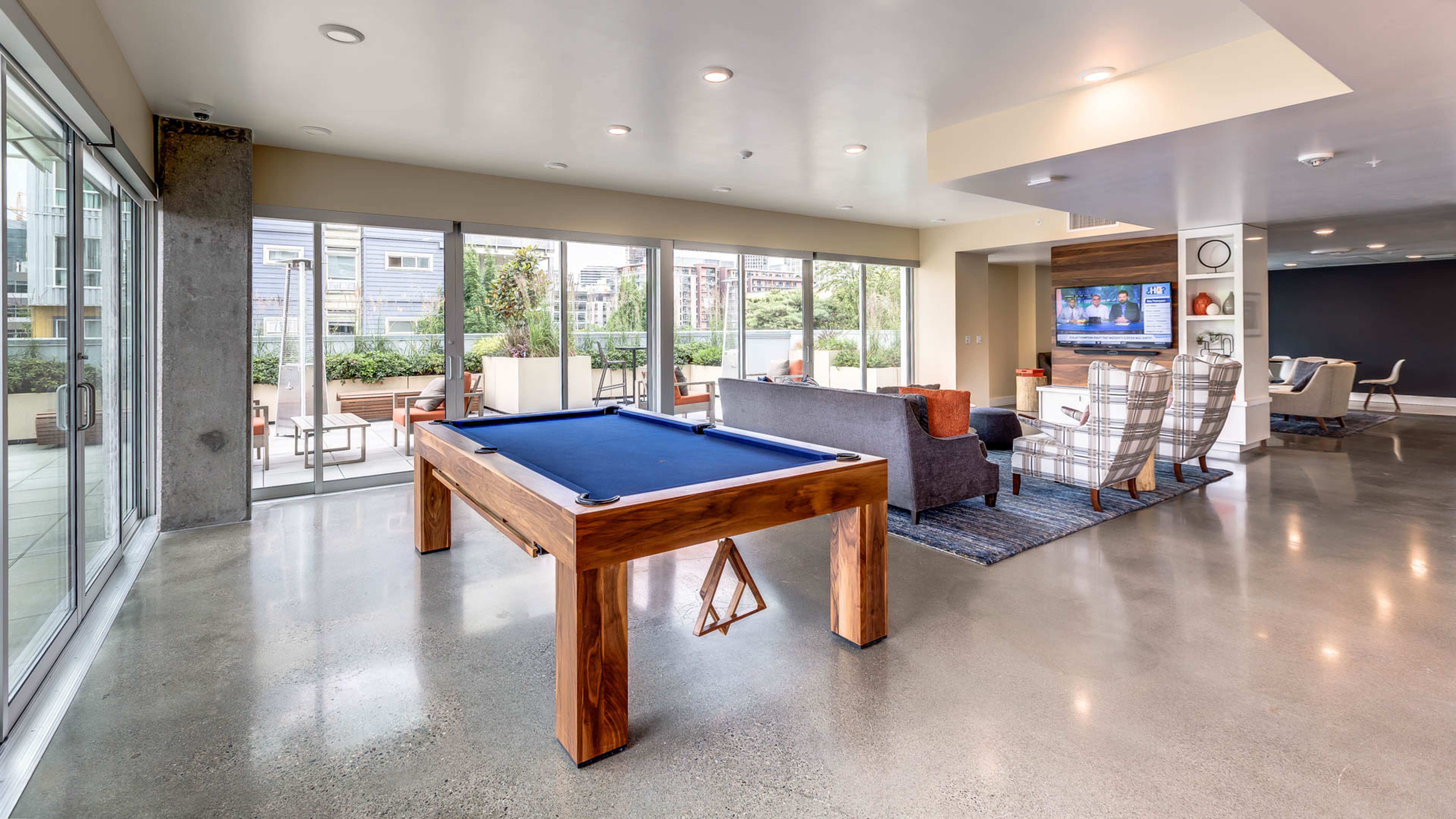 The image shows a modern indoor recreation area featuring a pool table, seating arrangements, large windows, and a television.