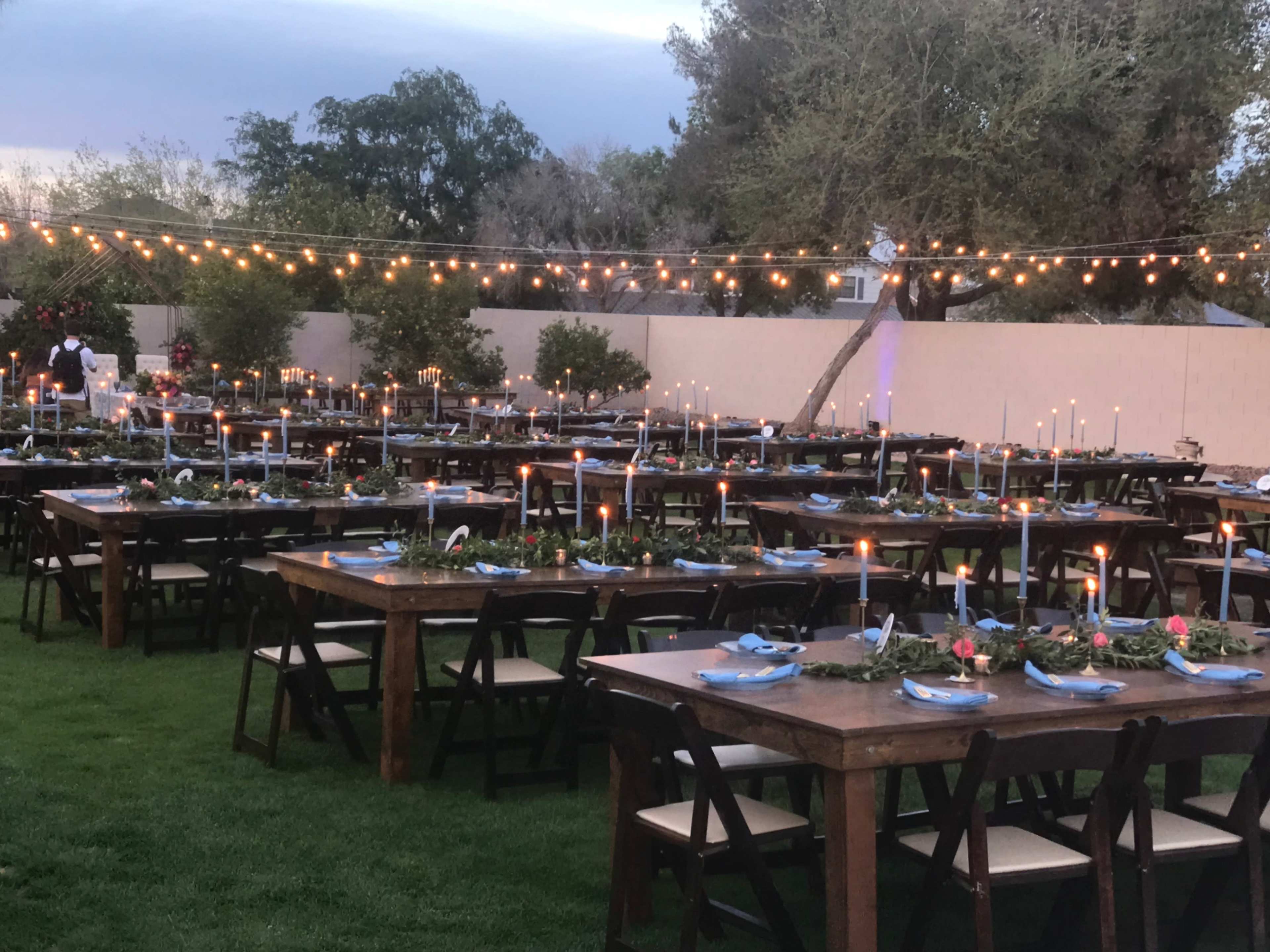A large outdoor dining setup features multiple wooden tables adorned with candles and decorative centerpieces, all illuminated by string lights overhead.