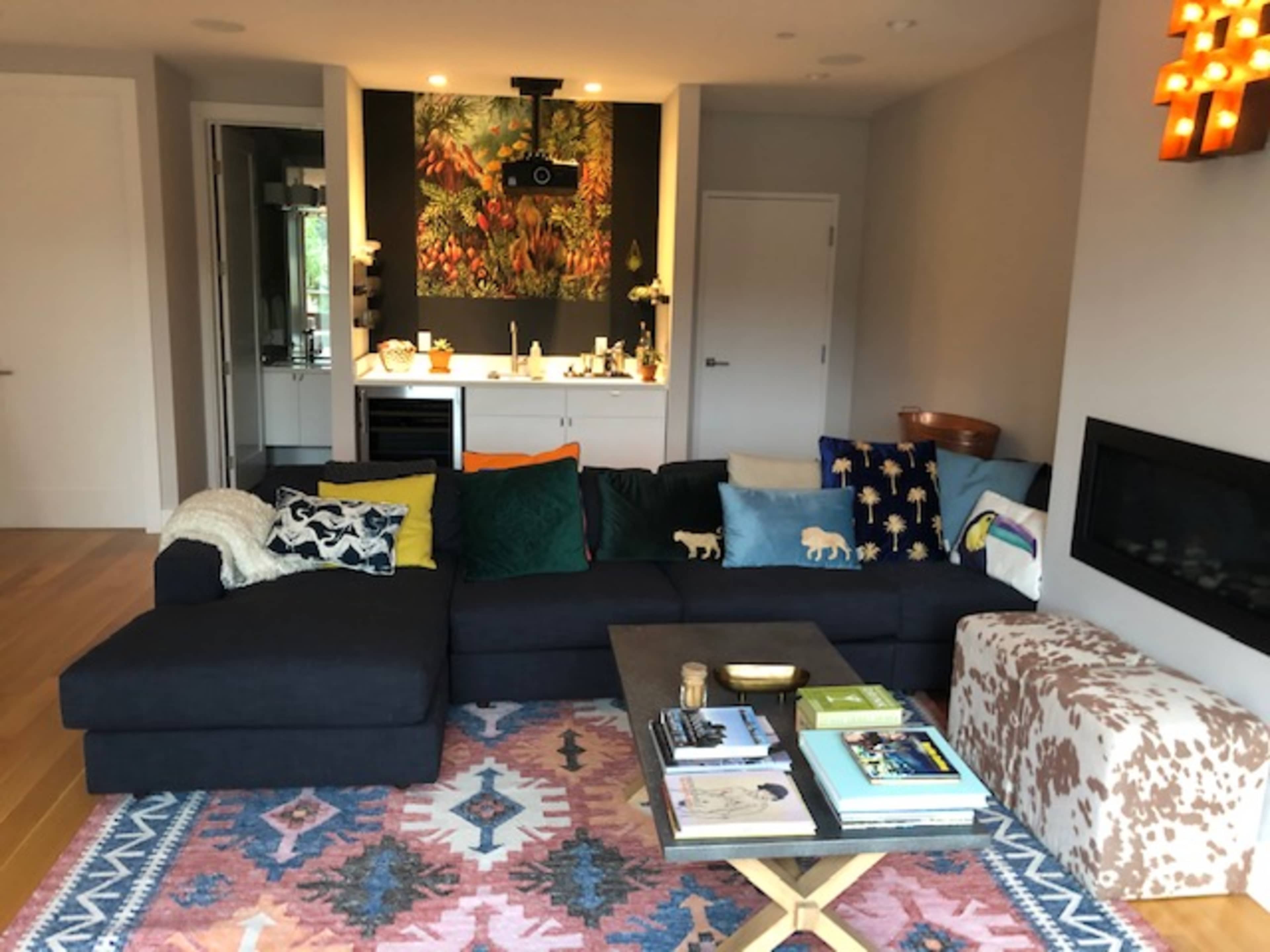 The image shows a modern living room featuring a dark sectional sofa, a patterned area rug, a wooden coffee table, and a wall adorned with a colorful artwork.