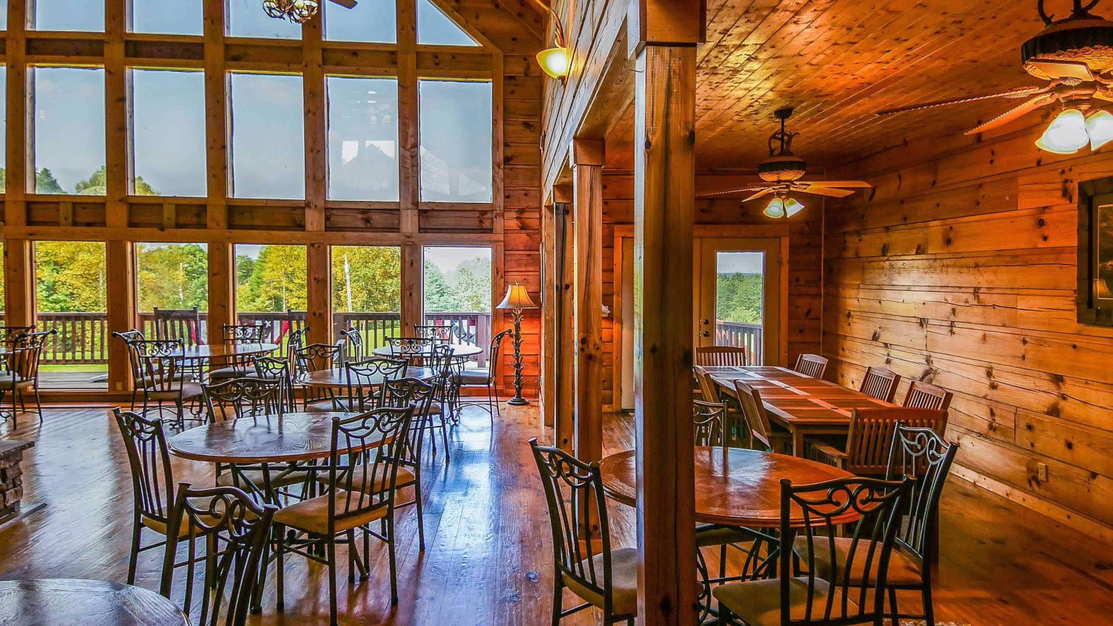 The image shows a spacious wooden interior of a restaurant with tables and chairs arranged near large windows overlooking a scenic view.