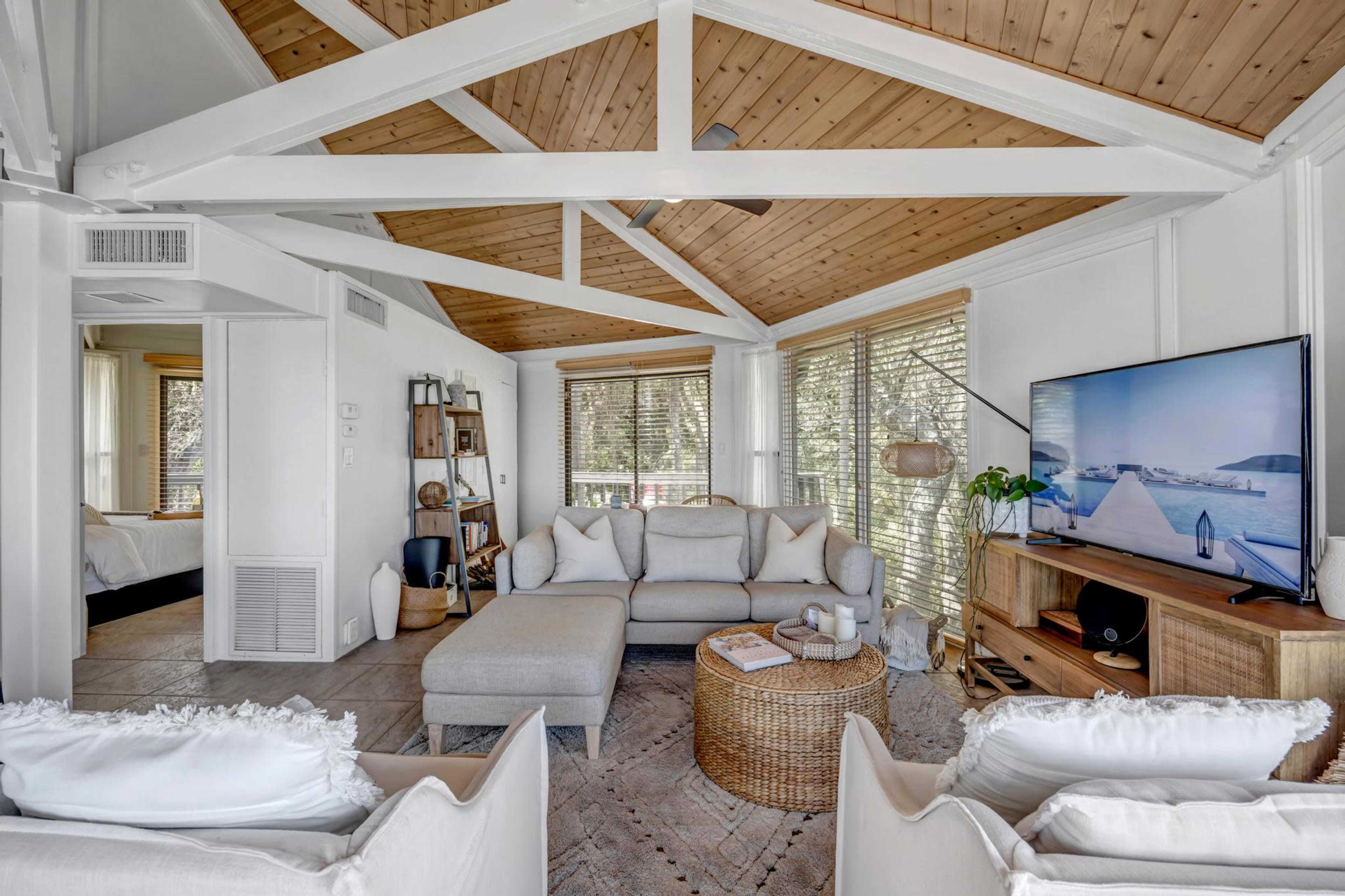 The living room features a large sectional sofa, a round wicker coffee table, and a flat-screen TV against a wall with exposed wooden beams overhead.