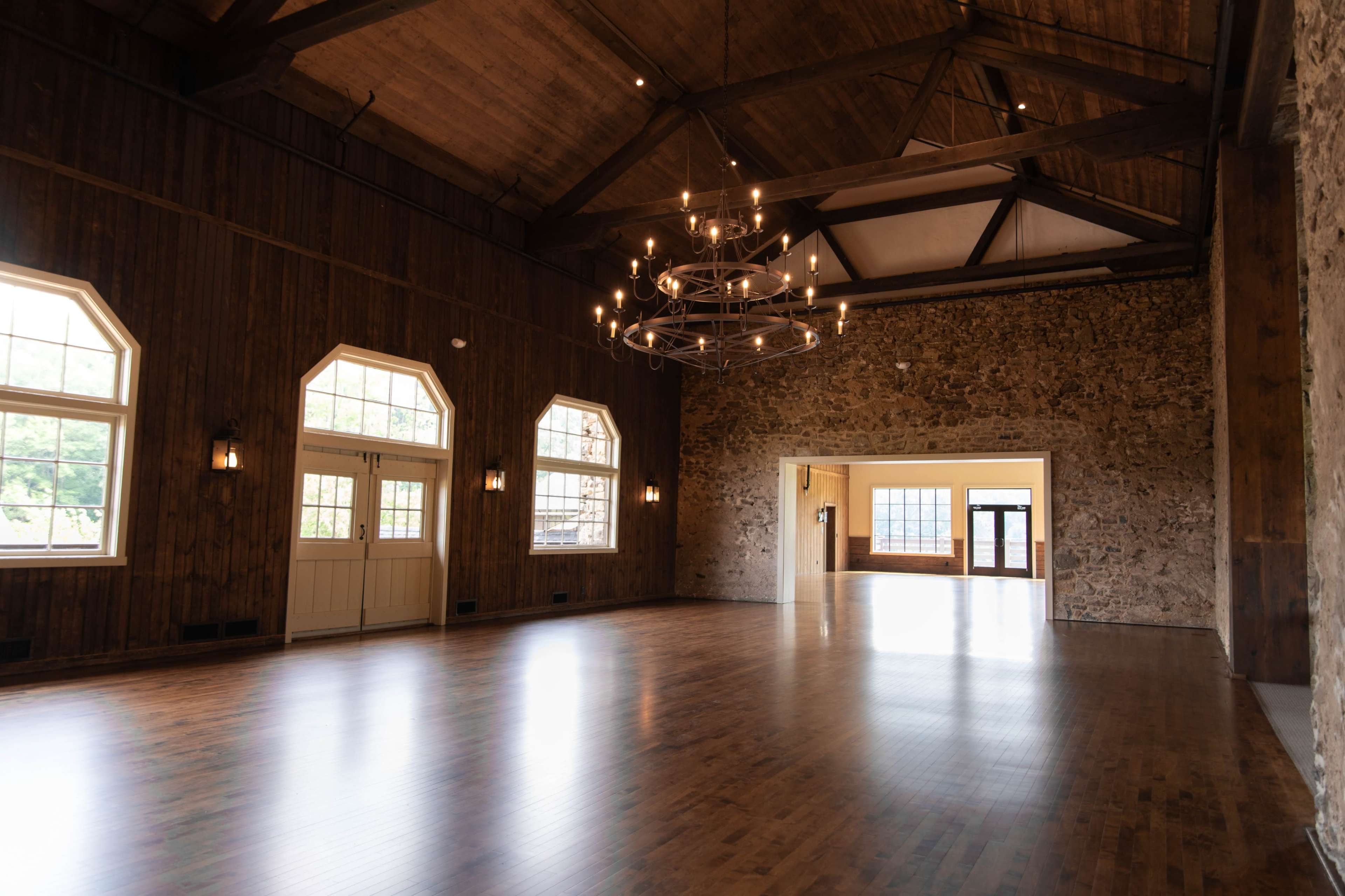 The image shows a spacious indoor hall with wooden beams, large windows, and a stone accent wall, featuring polished wooden floors and a modern chandelier.