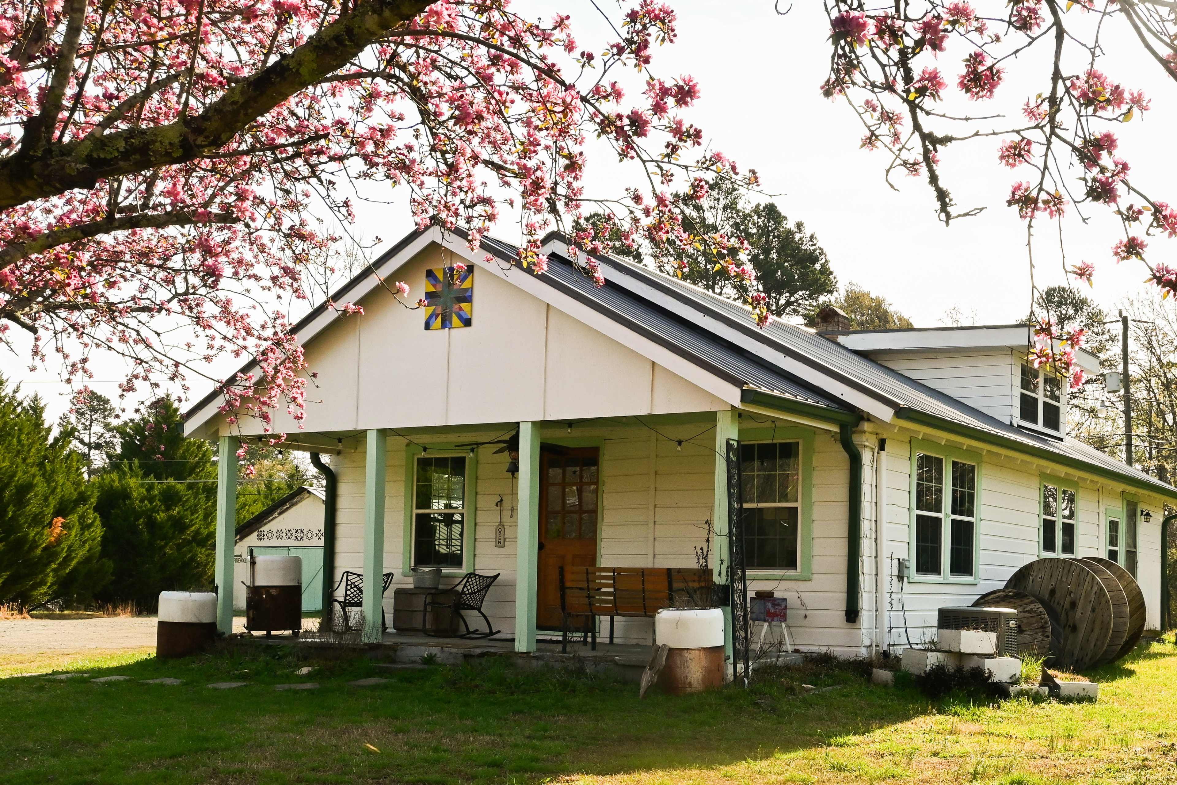 A white farmhouse with a black roof is surrounded by blooming pink trees and features a porch with wooden chairs and a decorative flower emblem.