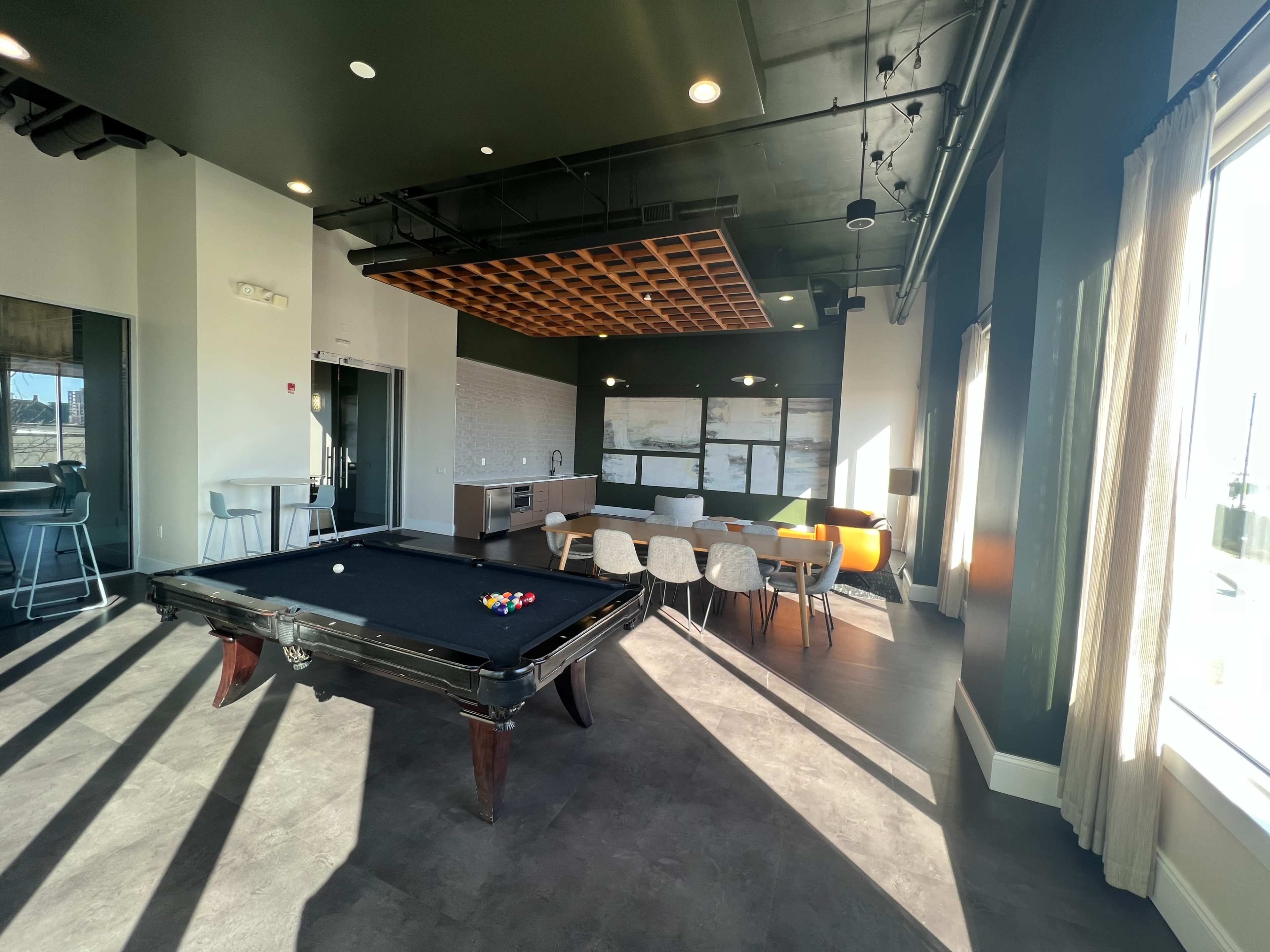 A modern recreation room featuring a billiard table, seating areas, and large windows allowing natural light.