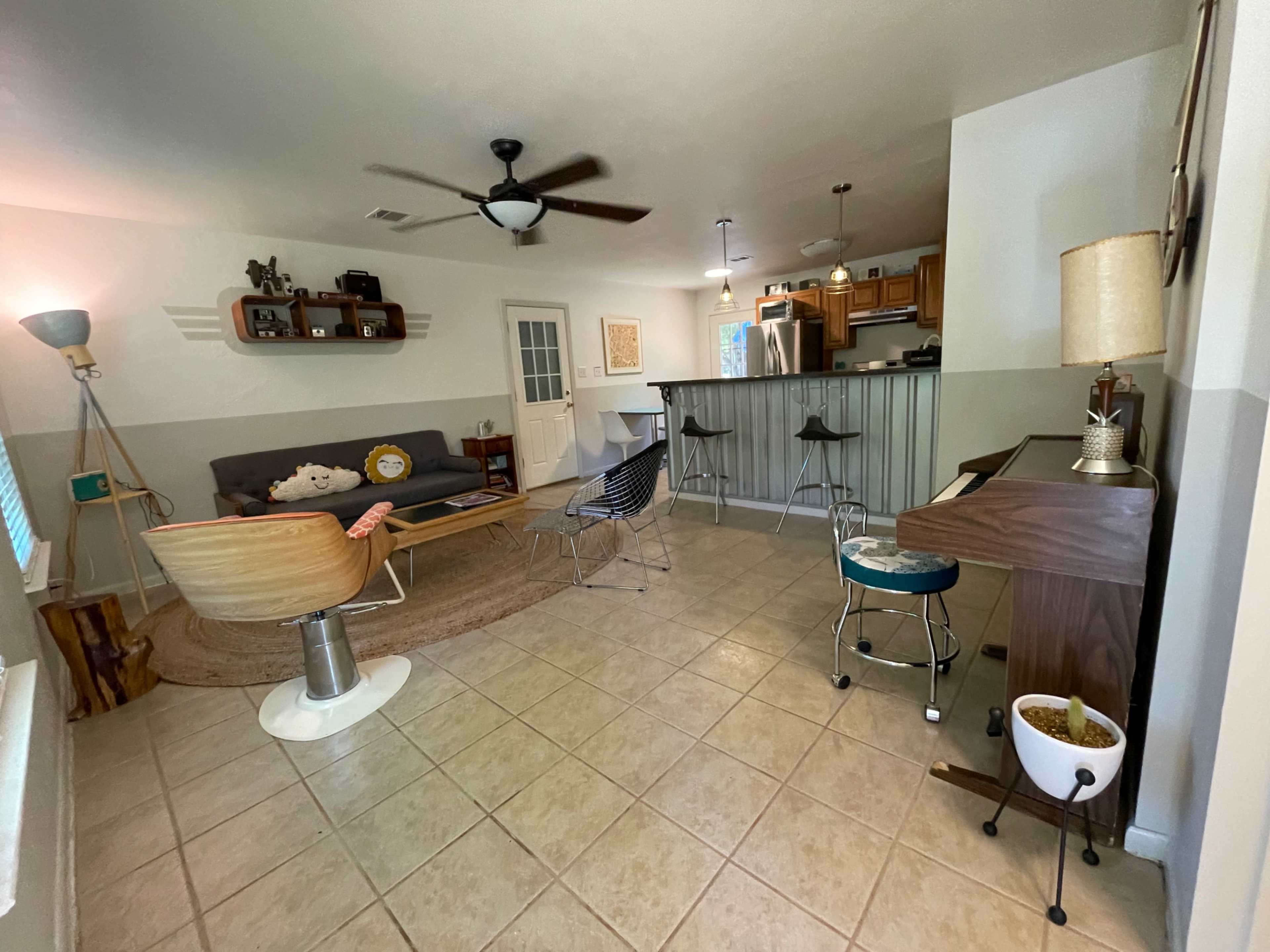 The image shows a spacious living area with a couch, armchairs, a coffee table, a small piano, and a kitchen bar in the background.