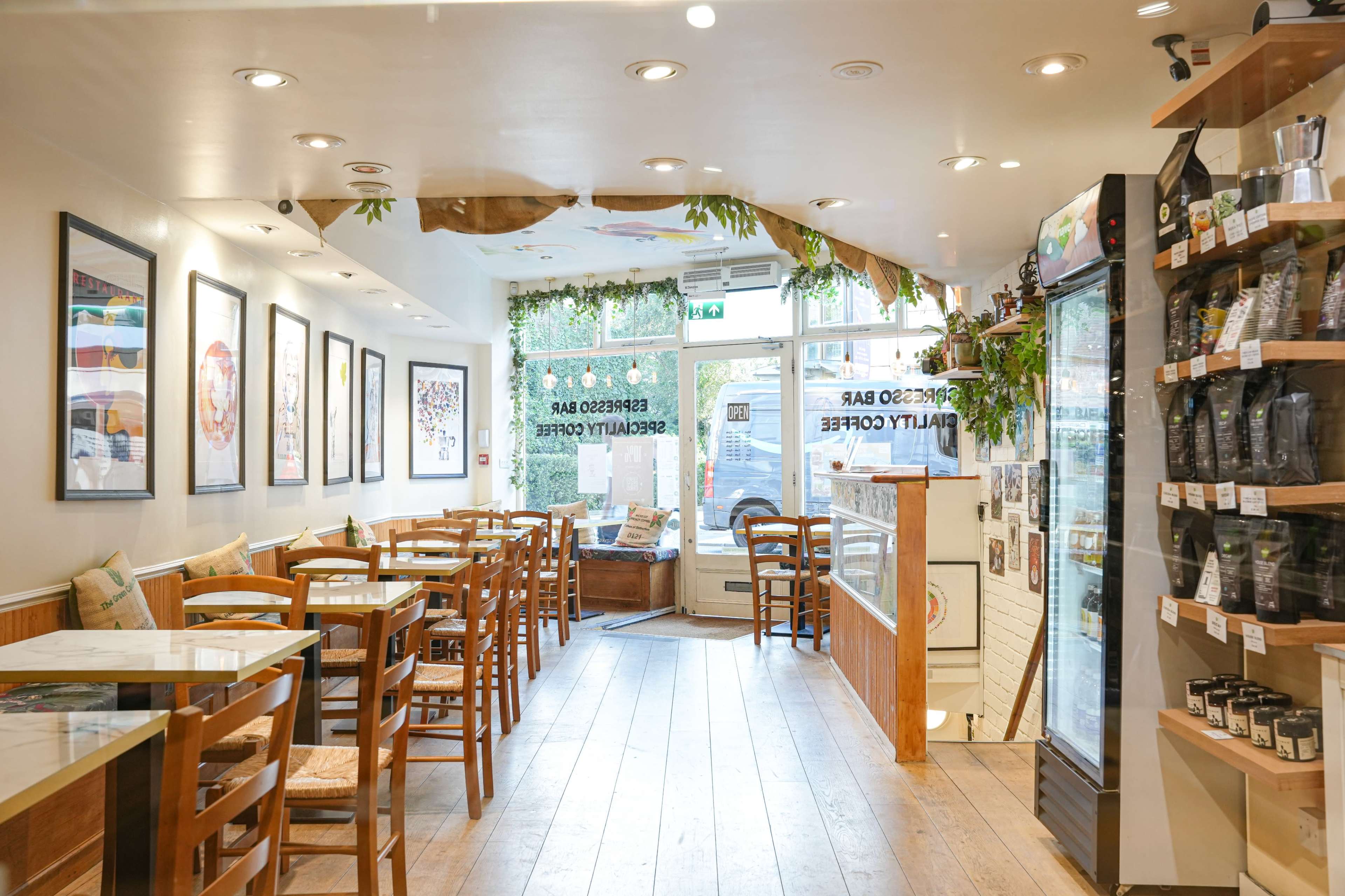 A cozy café features wooden tables and chairs, framed artwork on the walls, and a glass display case filled with coffee products.