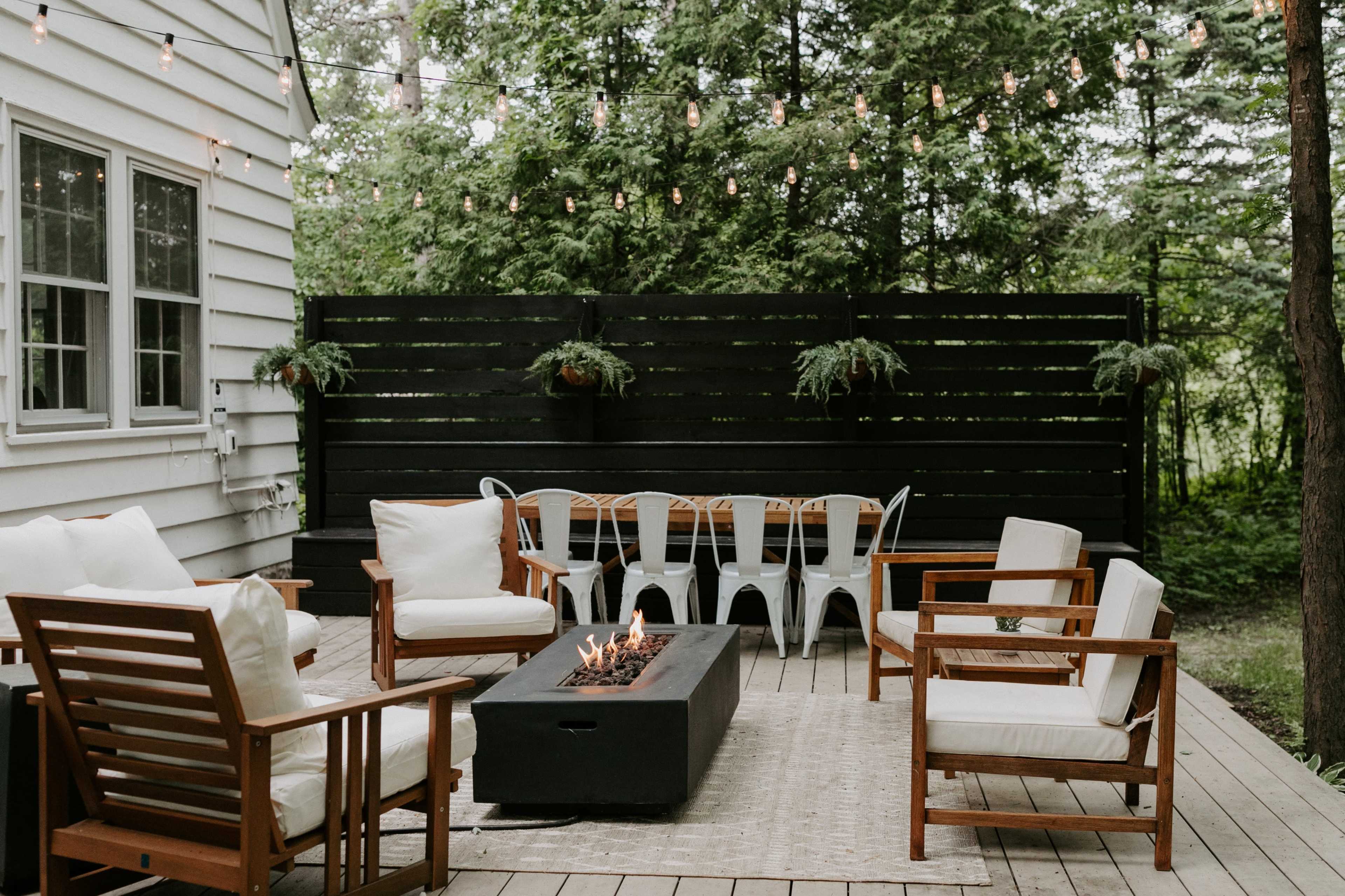 A comfortable outdoor patio features wooden furniture, a fire pit, and string lights surrounded by greenery.