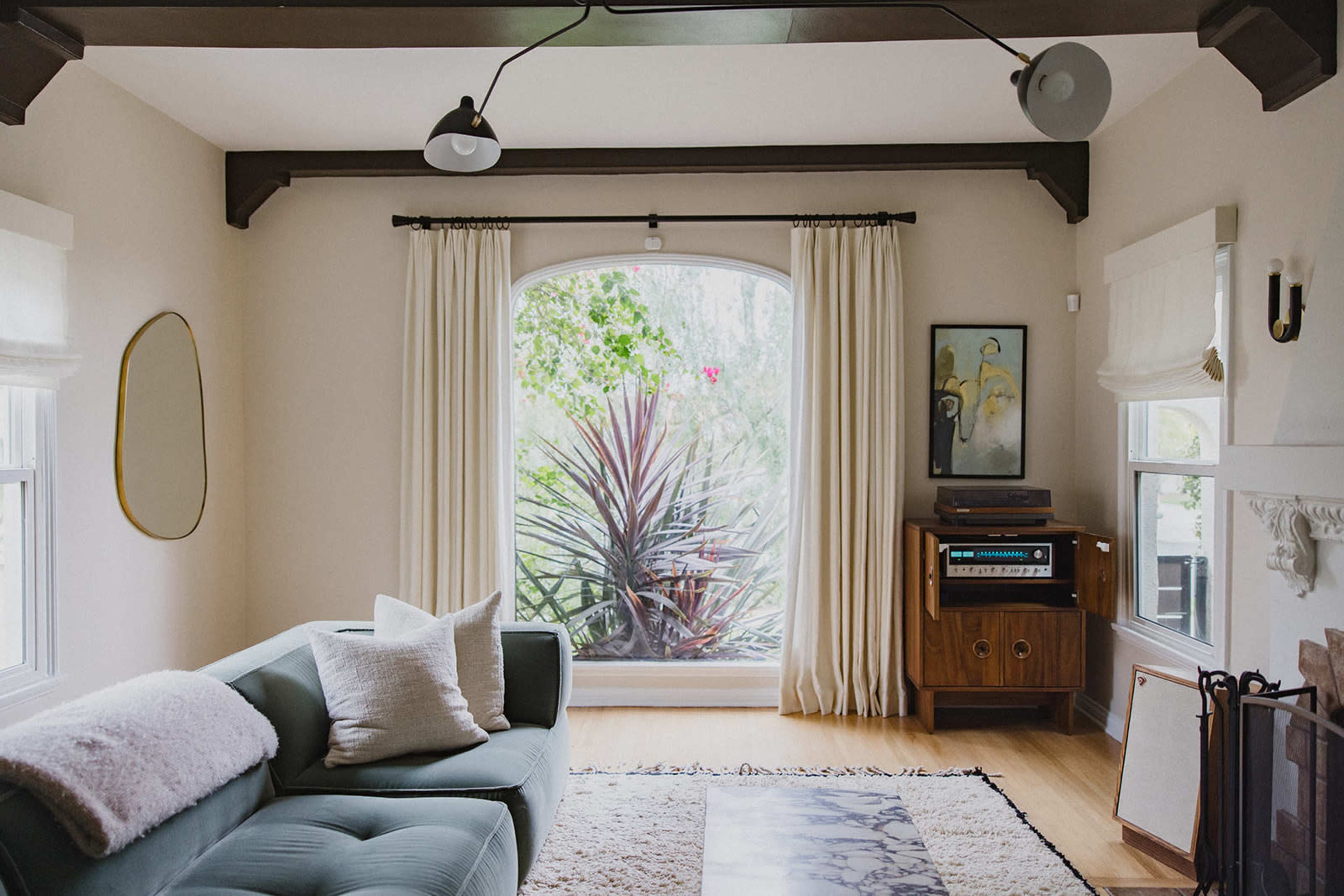 A light-filled living room features a green sectional sofa, large windows with curtains, and a vintage stereo unit beside a wooden cabinet.