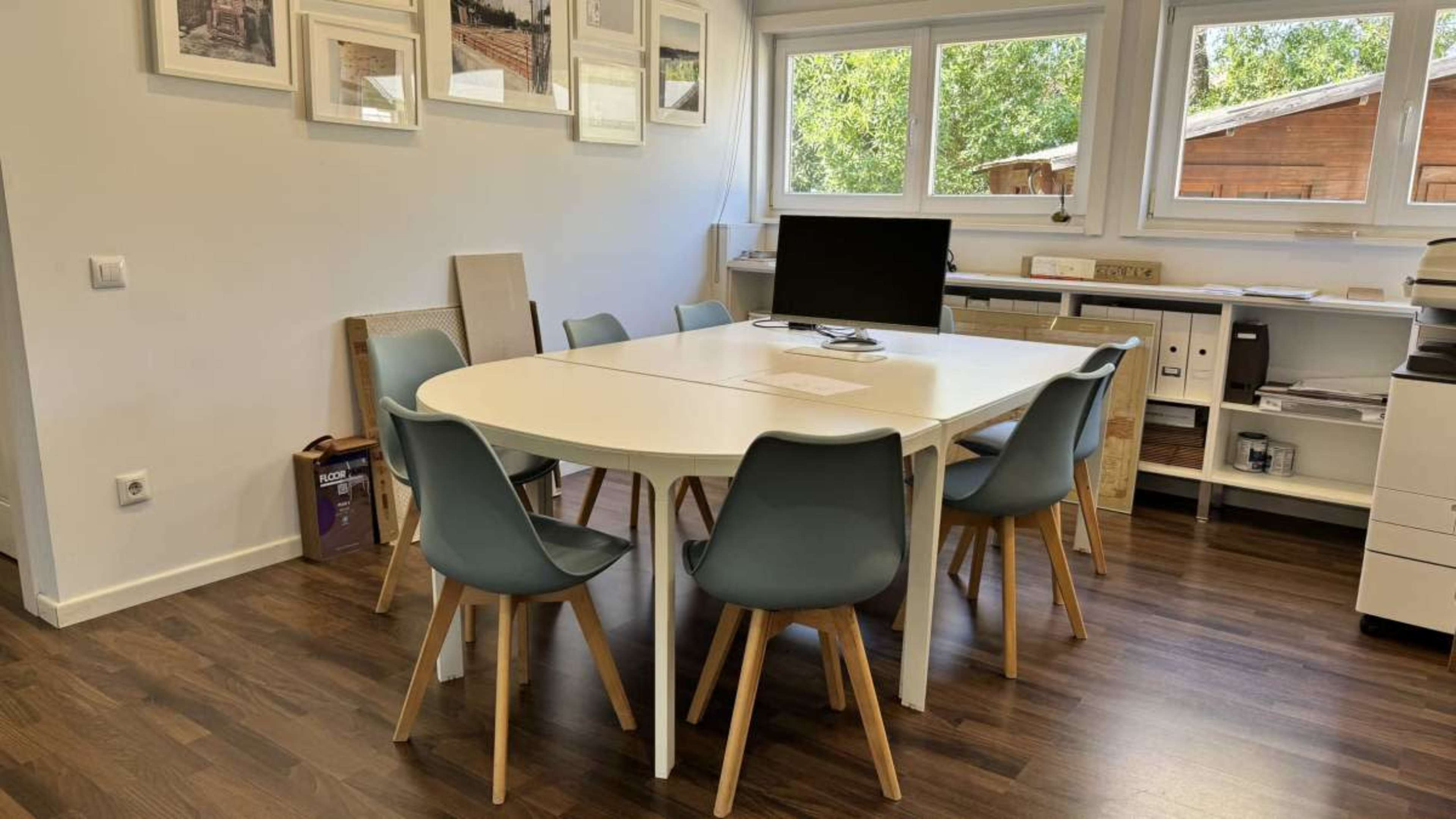 The image shows a modern meeting room with a round white table surrounded by six blue chairs, a computer monitor, and several framed pictures on the wall.