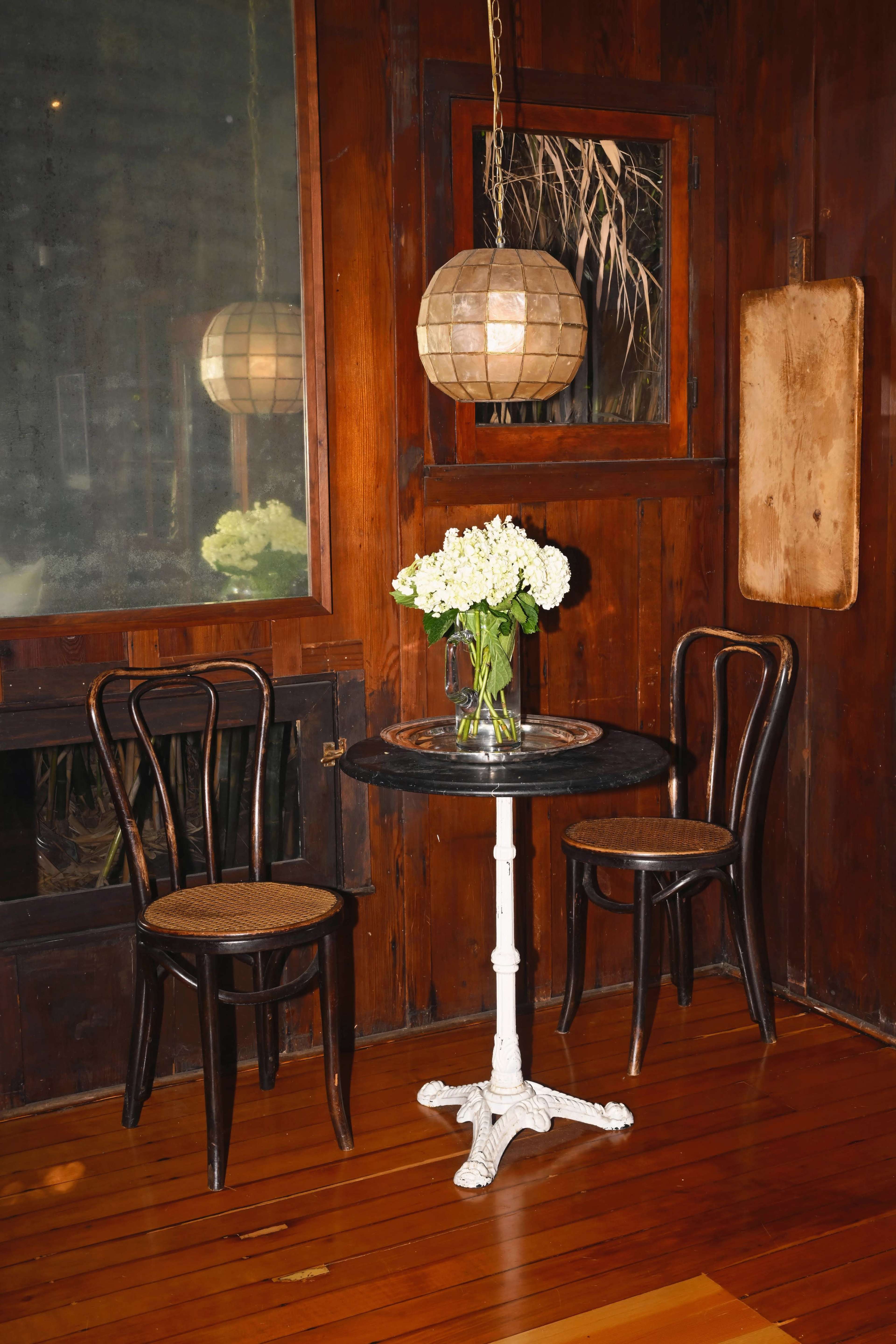 A small round table with a floral arrangement sits between two wooden chairs in a dimly lit, wooden-paneled room.