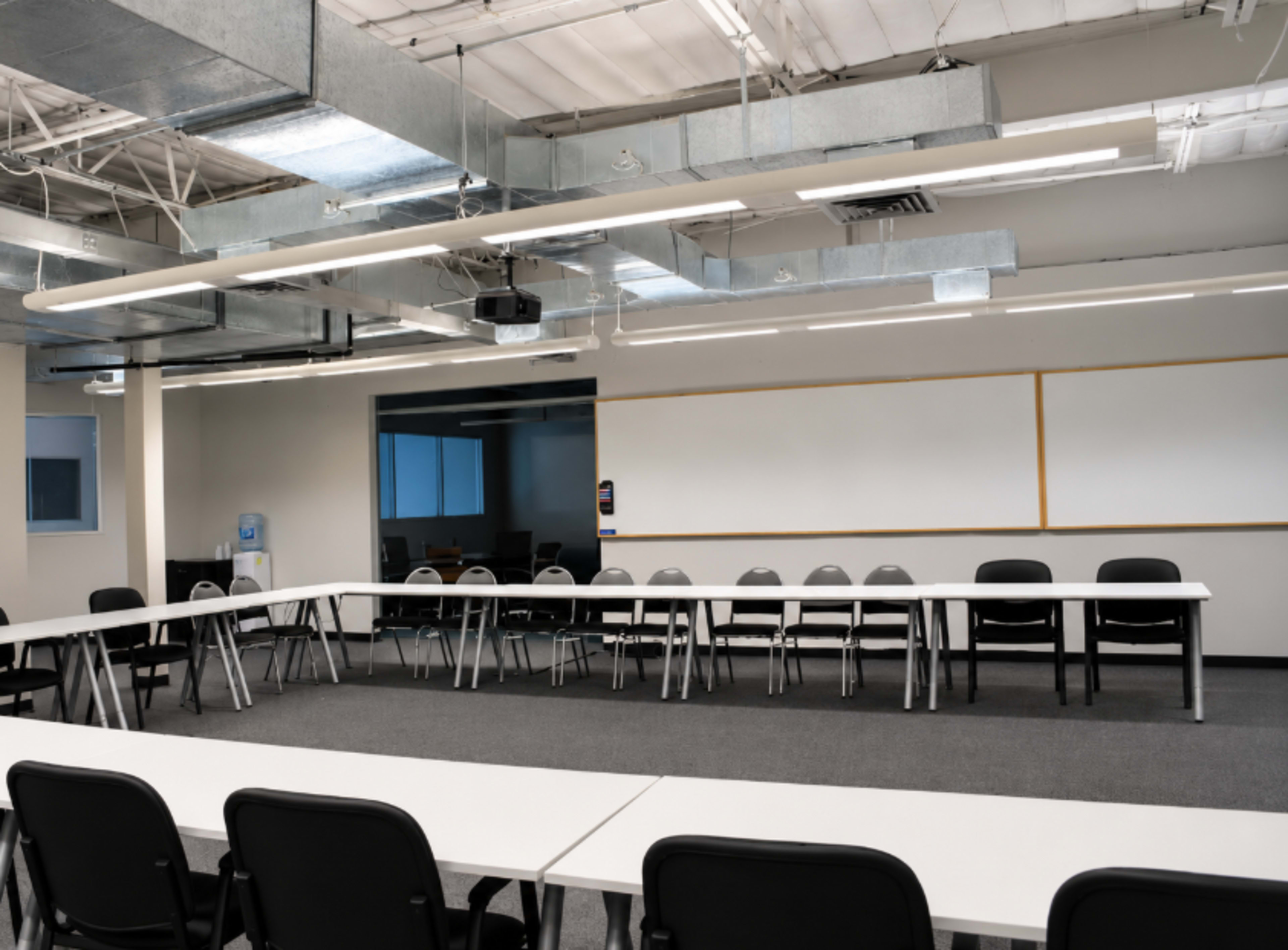 A spacious meeting room features a large rectangular table surrounded by chairs, with walls equipped for presentations and natural light coming from windows.