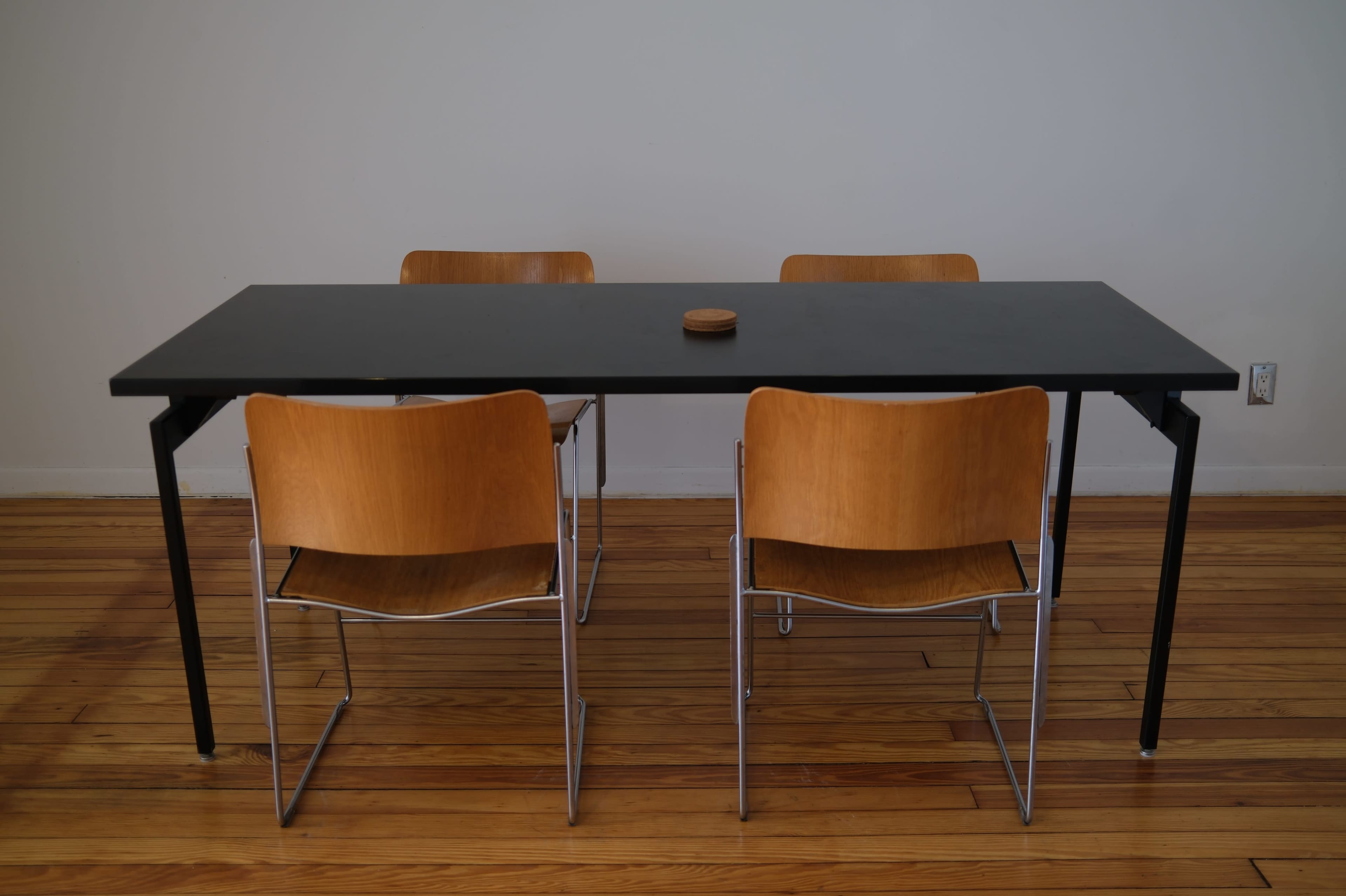 A rectangular black table is surrounded by four wooden chairs on a wooden floor.