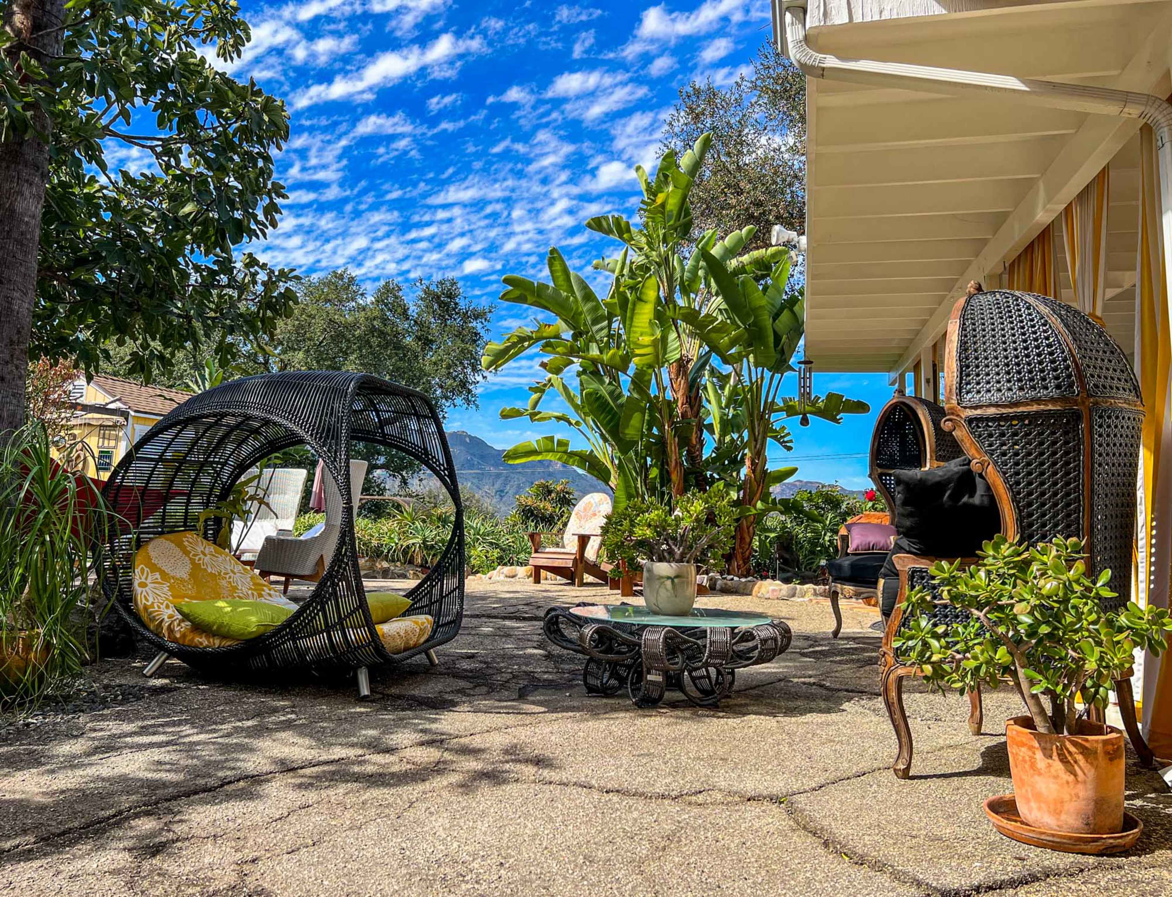 The image shows a scenic outdoor patio featuring two modern wicker chairs, a circular coffee table, and lush greenery under a blue sky with scattered clouds.