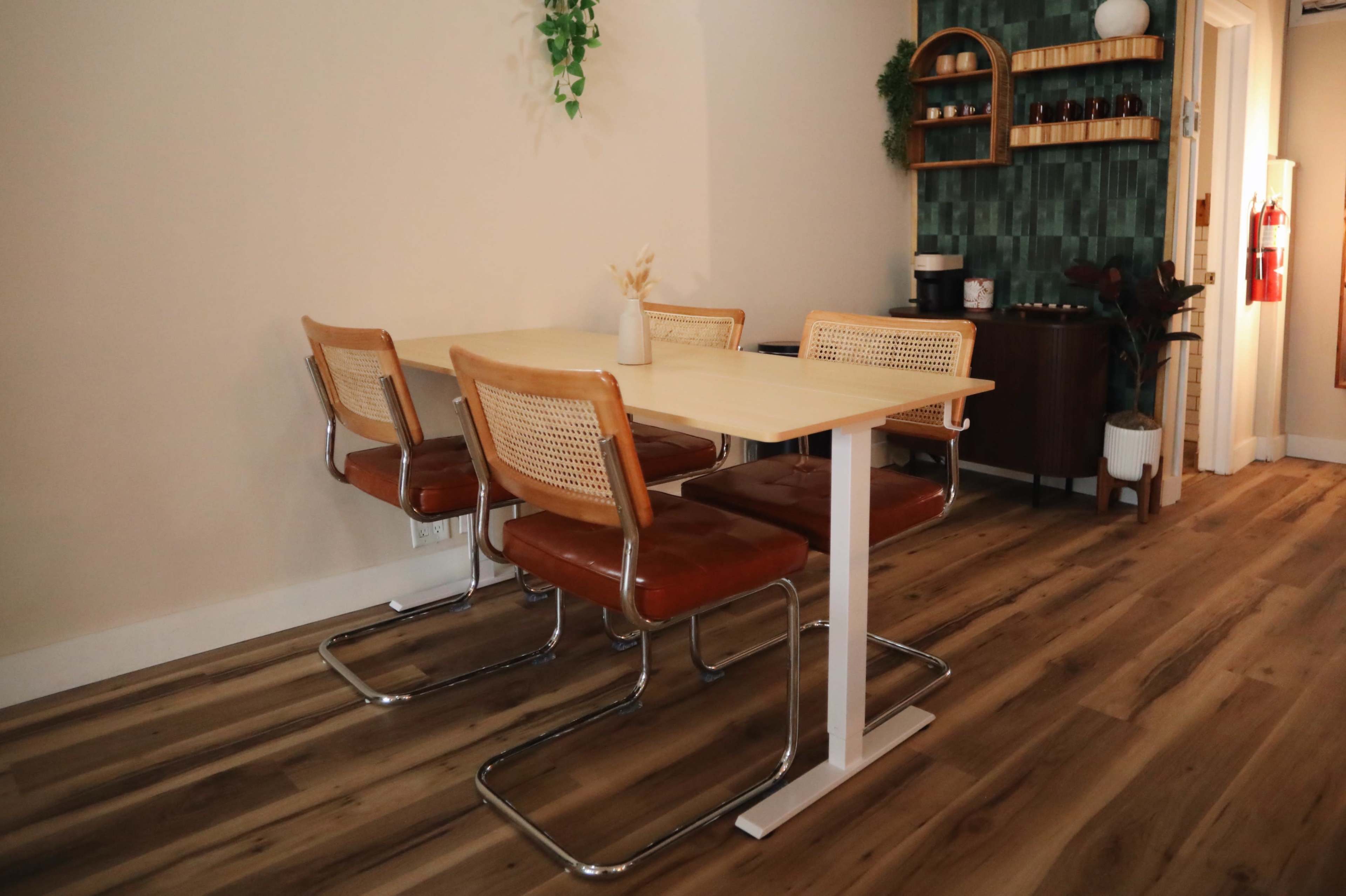 A modern dining area features a light-colored table surrounded by four brown and cream chairs, with a plant hanging above and a shelf in the background displaying kitchen items.