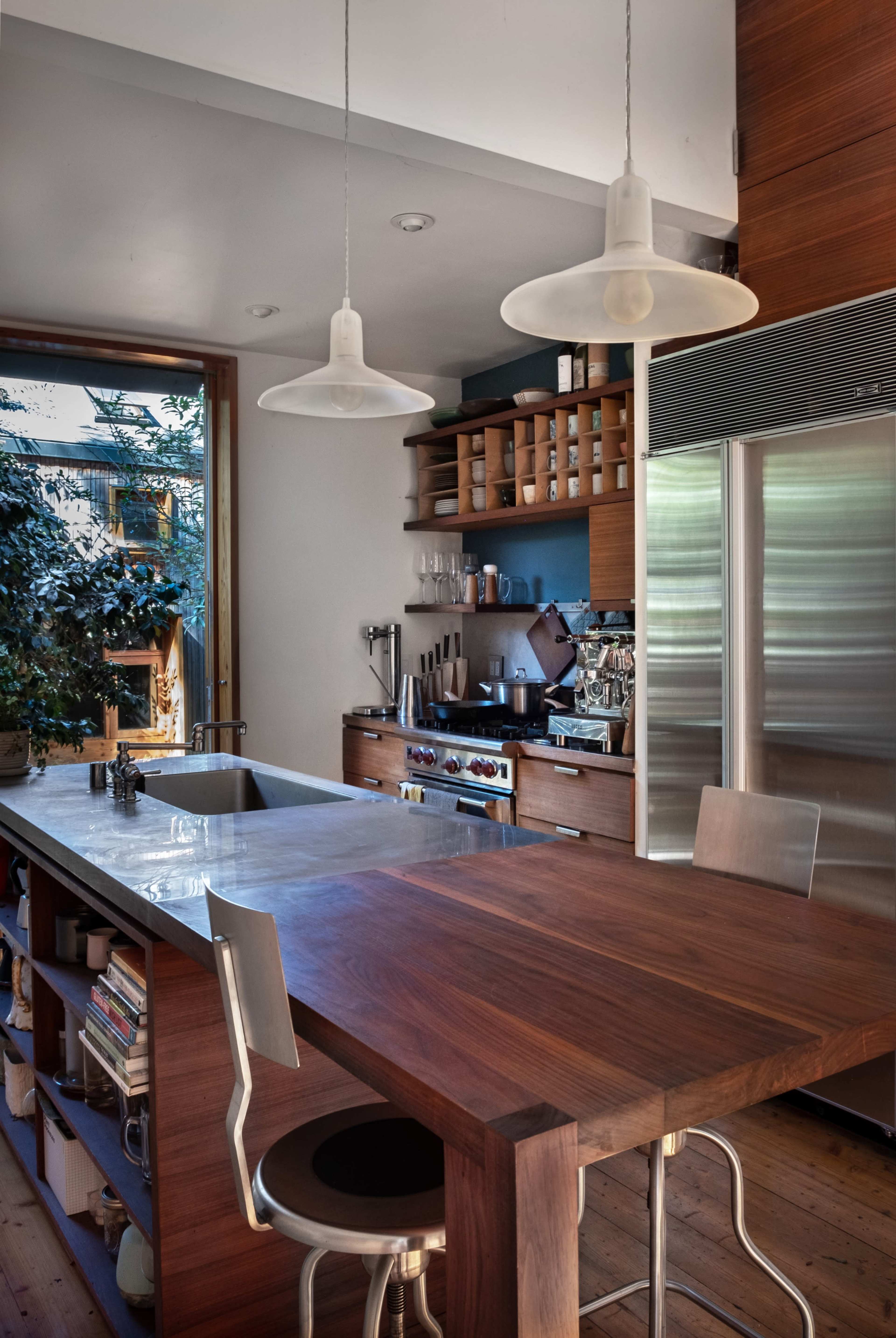 A modern kitchen features a large wooden island with metal bar stools, pendant lights hanging above, and open shelving stocked with various kitchen items.