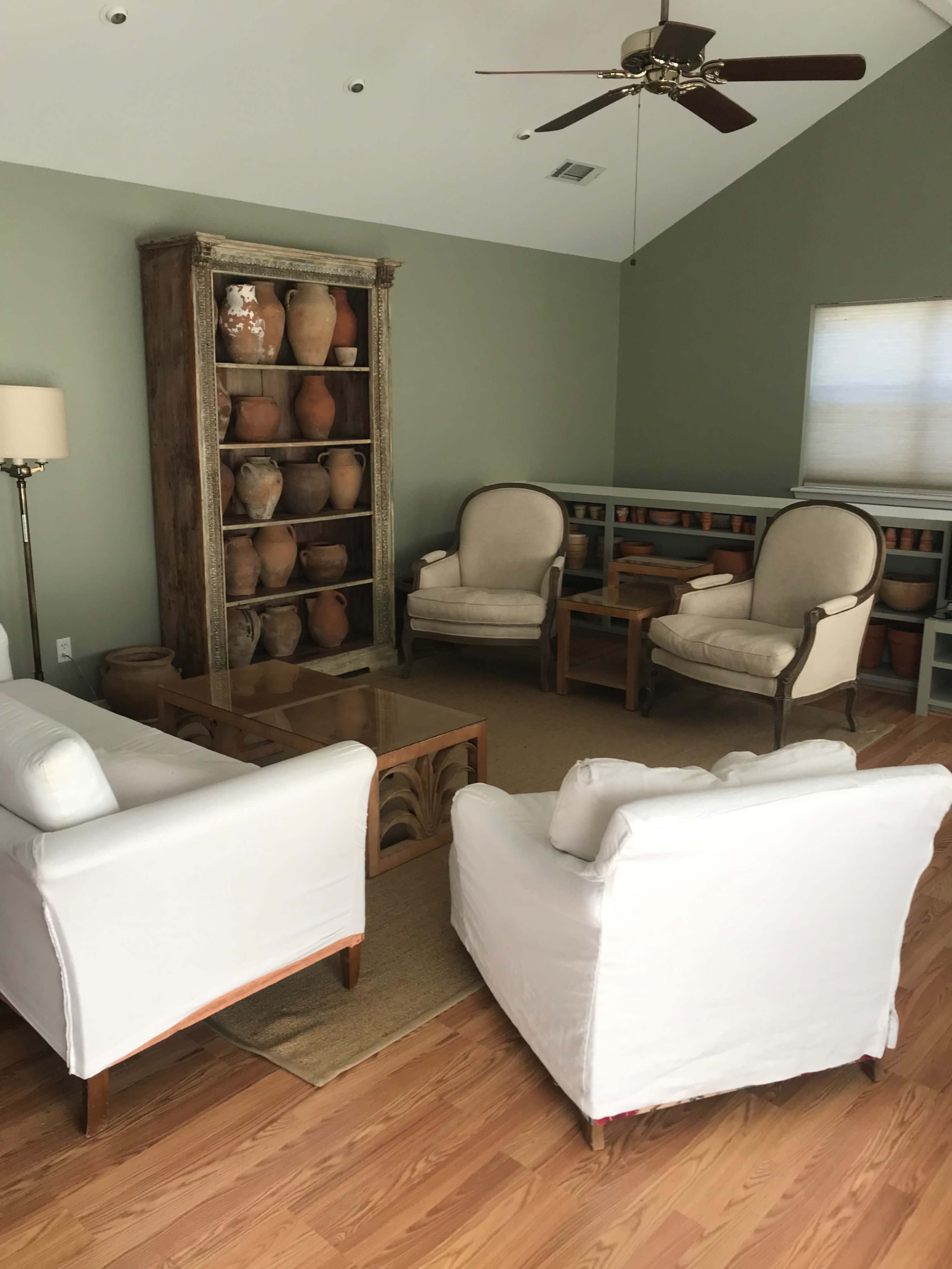 A living room with white upholstered chairs, a wooden coffee table, and a large display shelf filled with clay pots against a light green wall.