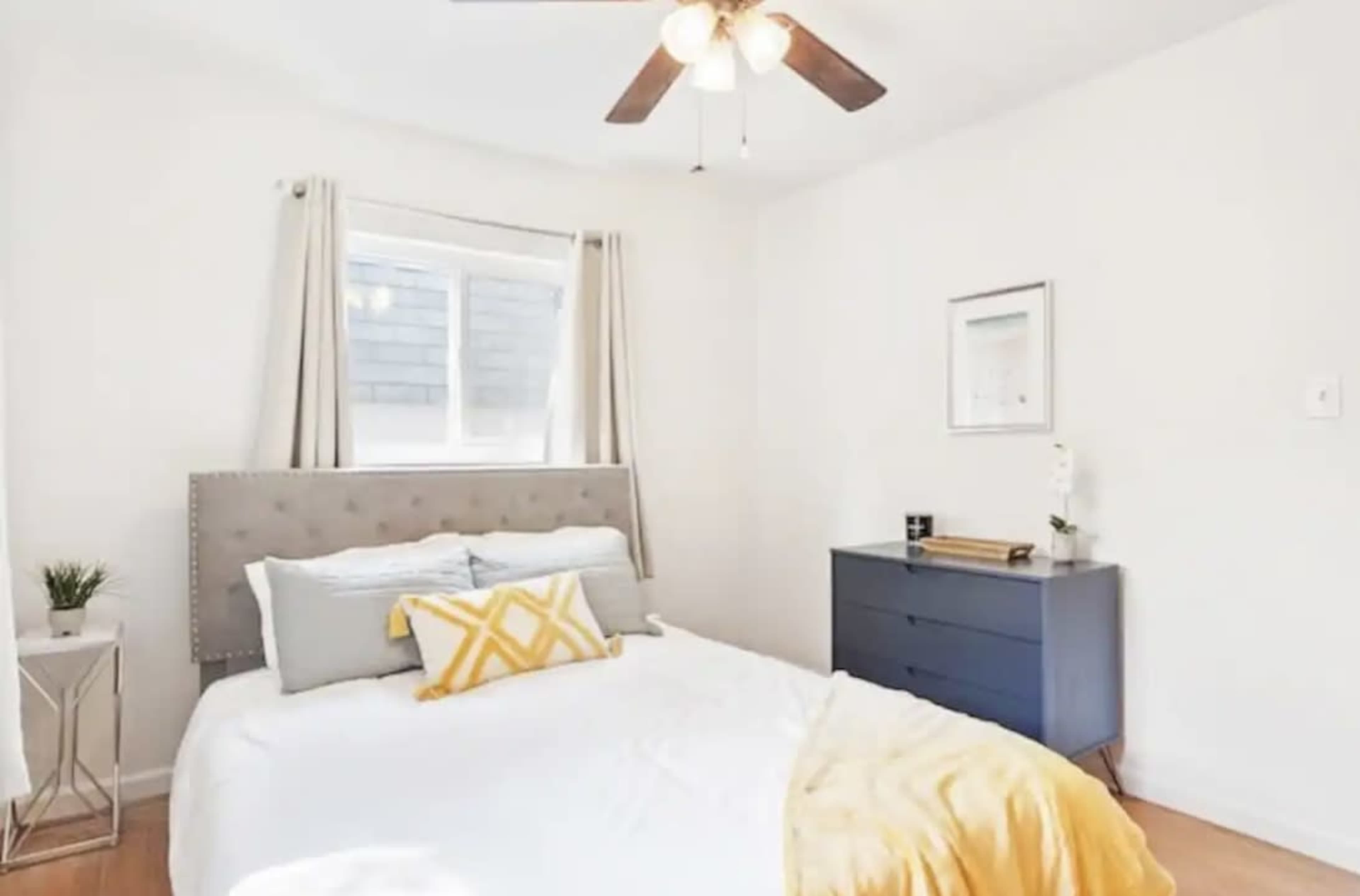 A bright bedroom features a queen-sized bed with a gray headboard, yellow and white accents, and a dark blue dresser against the wall.