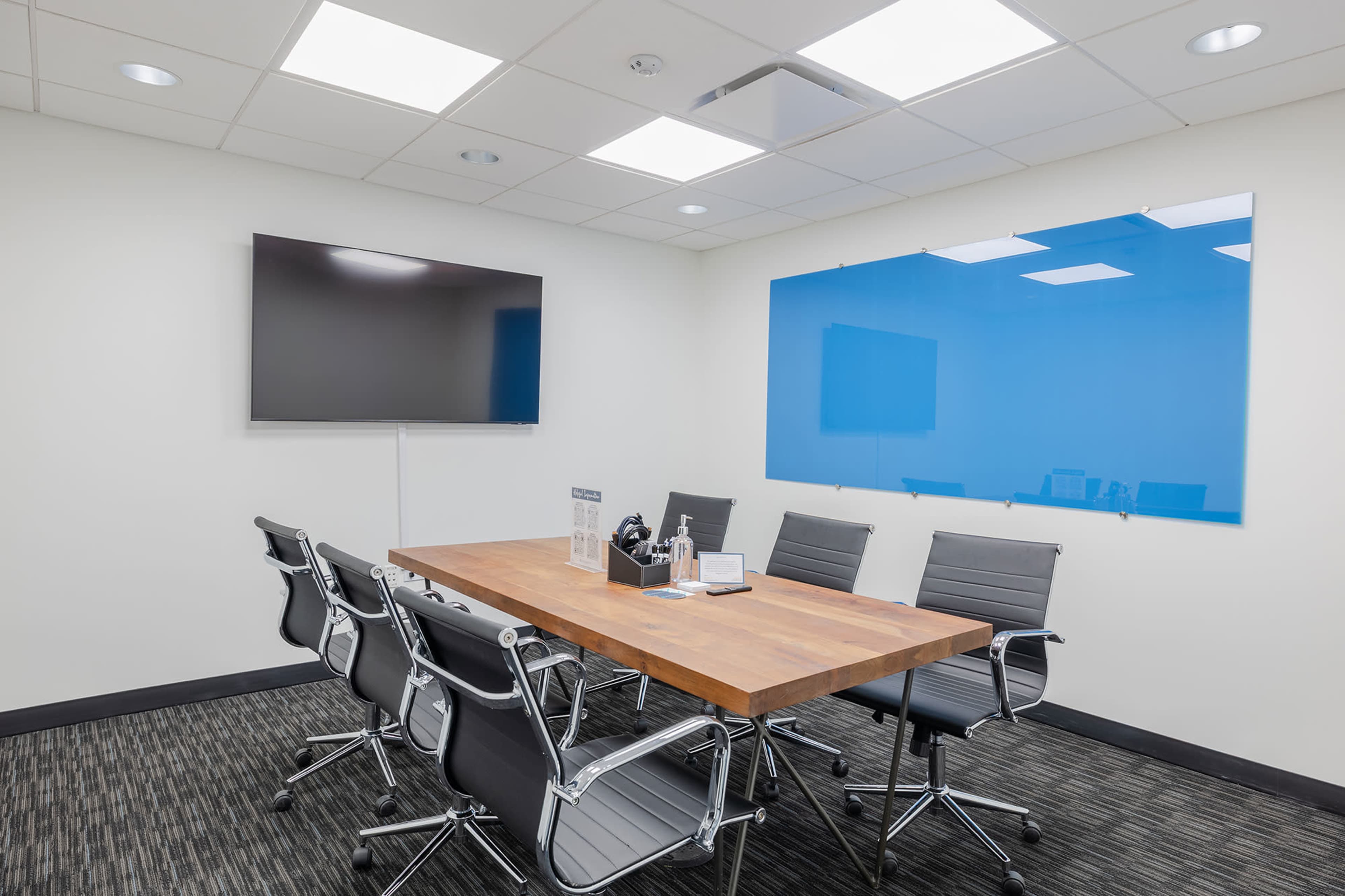 The image shows a conference room with a wooden meeting table, six black leather chairs, a large screen on the wall, and a blue glass whiteboard.
