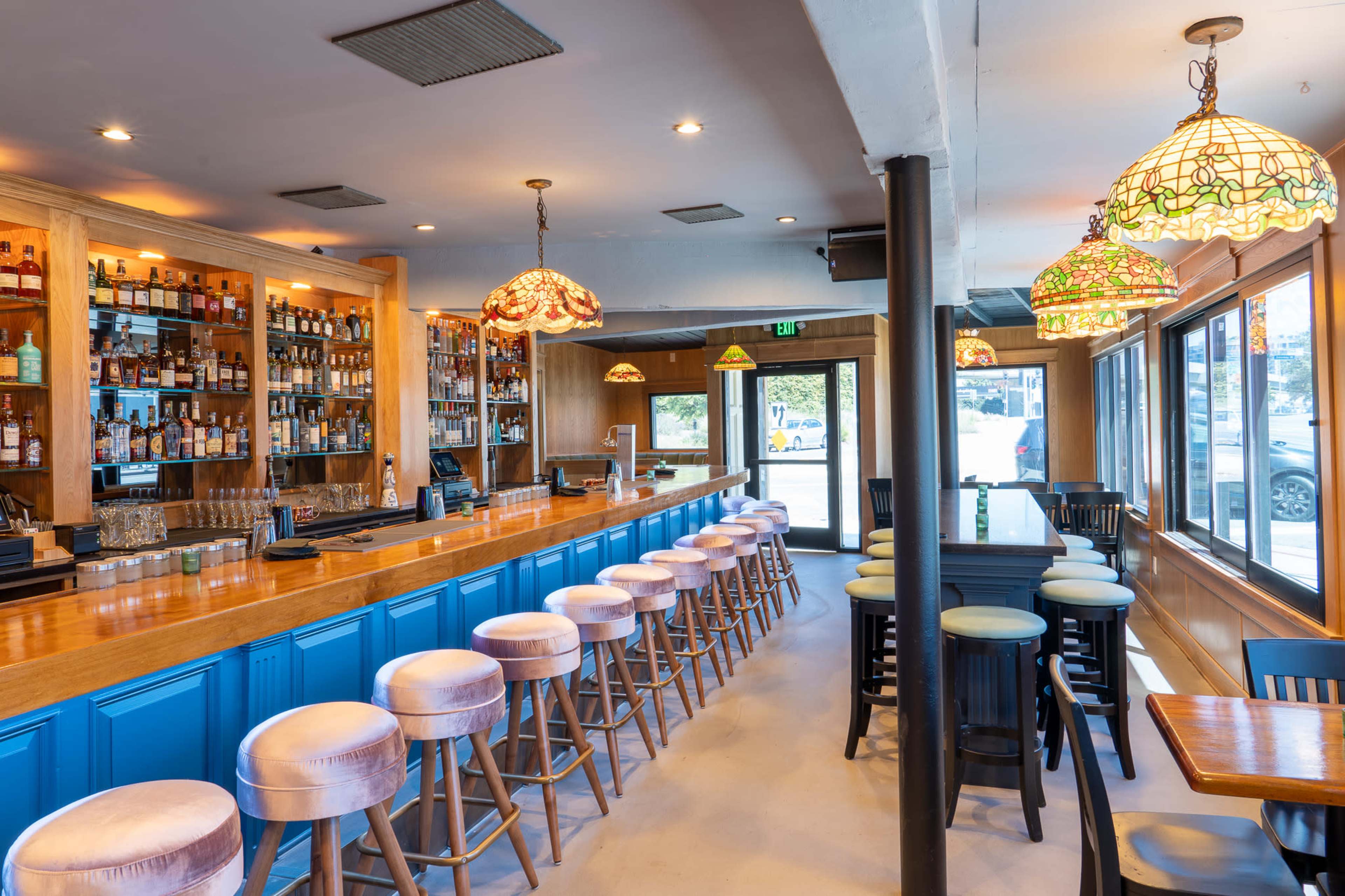 A spacious bar area with a wooden counter, high stools, and shelves lined with various bottles, illuminated by decorative pendant lights.