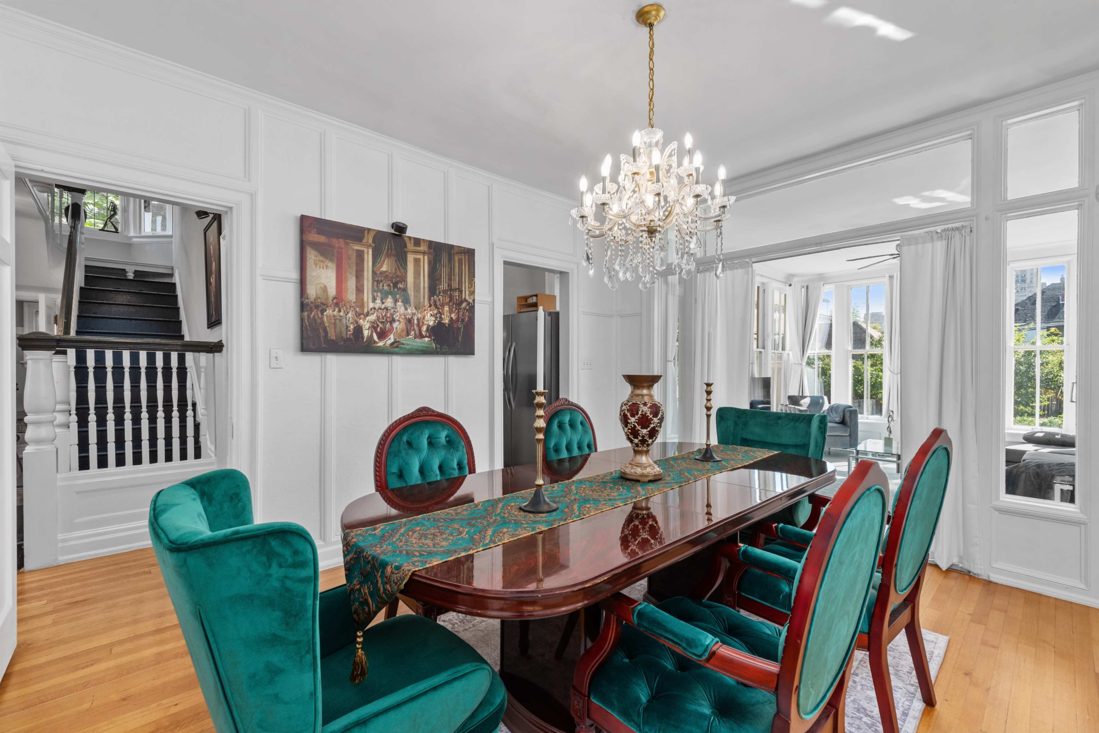 A dining room features a polished wooden table surrounded by green velvet chairs and a chandelier overhead, with large windows allowing natural light to enter.