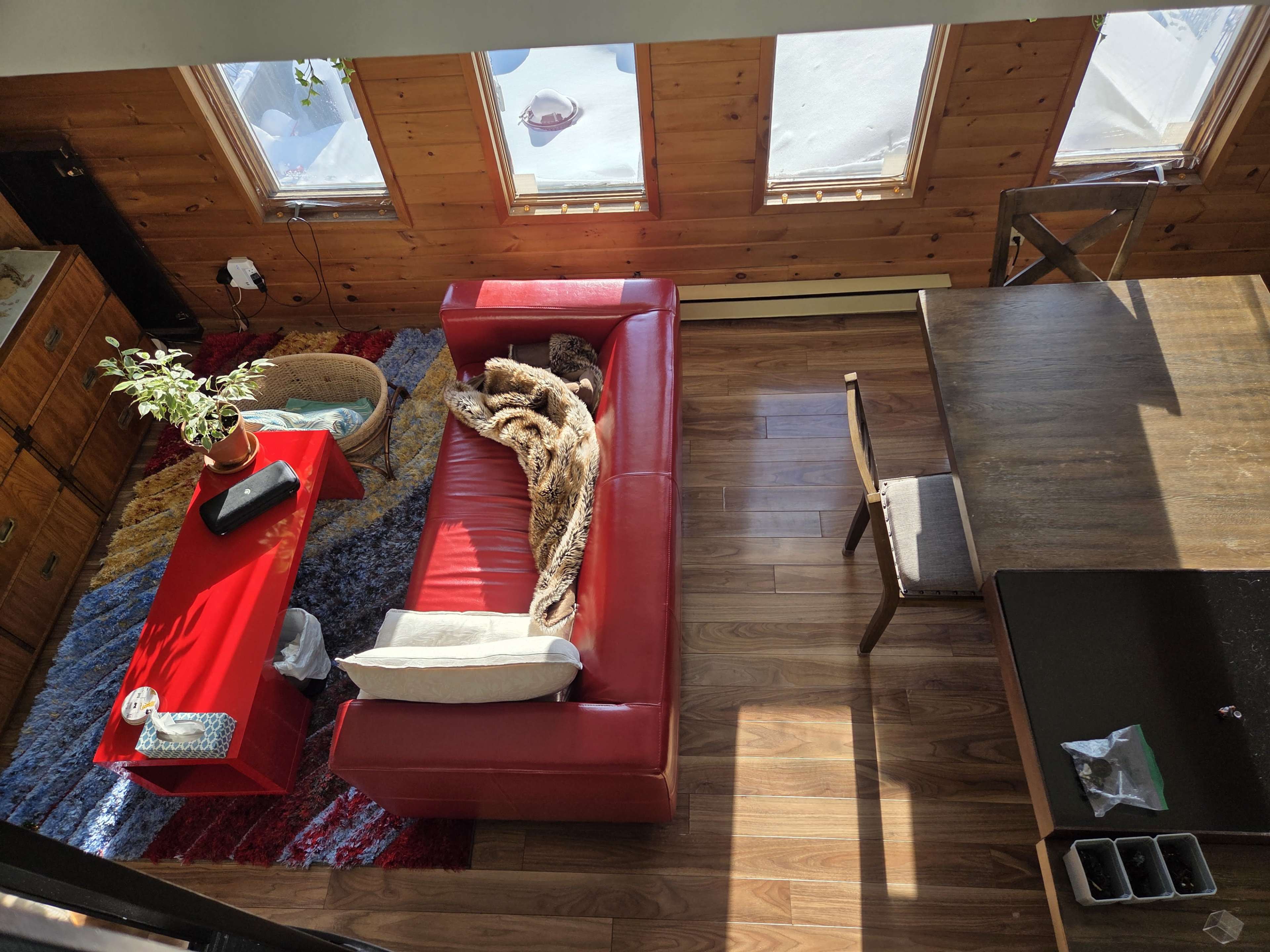 The image shows a cozy living room with a red sofa, coffee table, and a dining area, all surrounded by wooden walls and large windows letting in natural light.