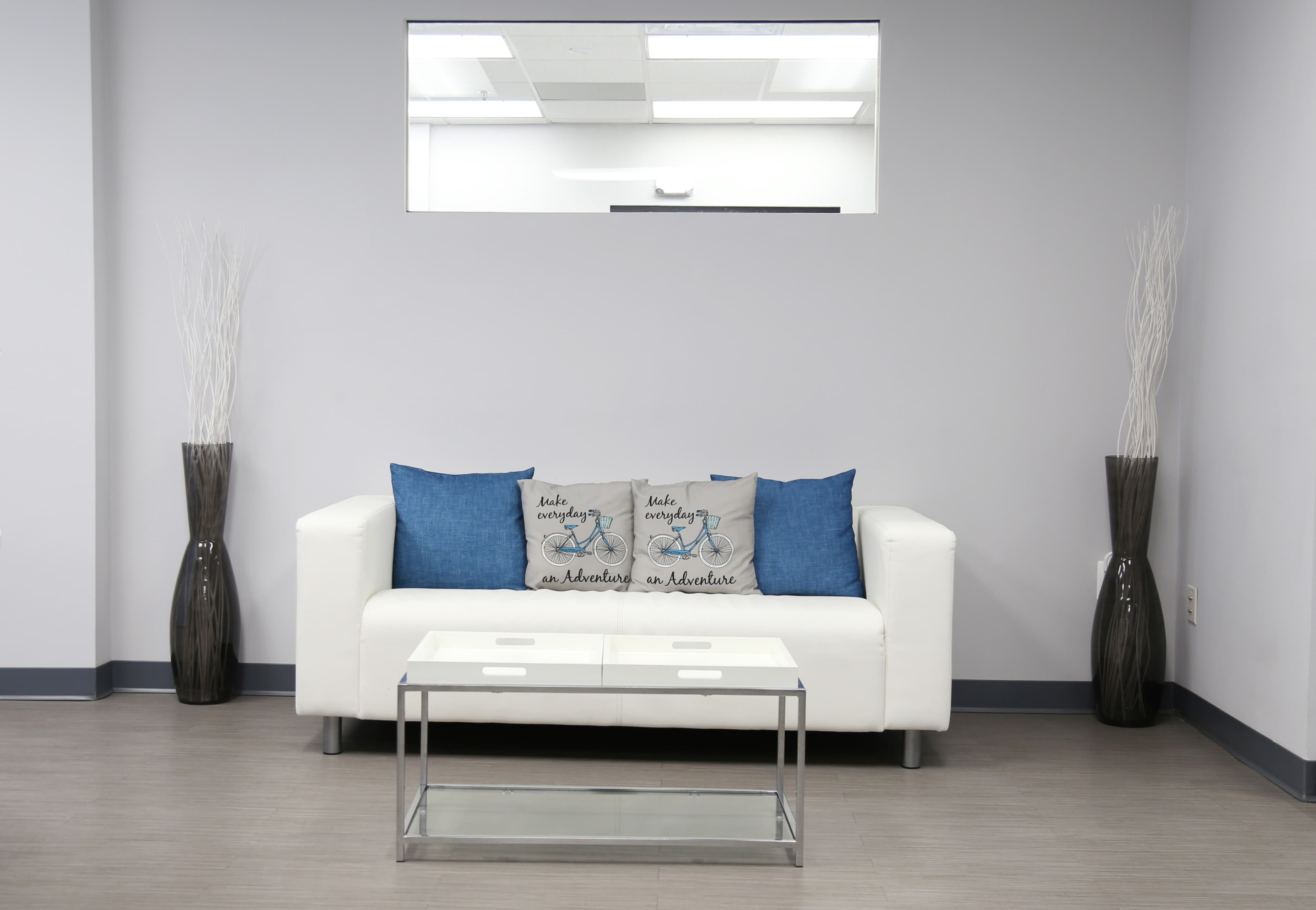 A modern living area features a white sofa with blue and gray cushions, a glass-topped coffee table, and tall decorative vases on either side.