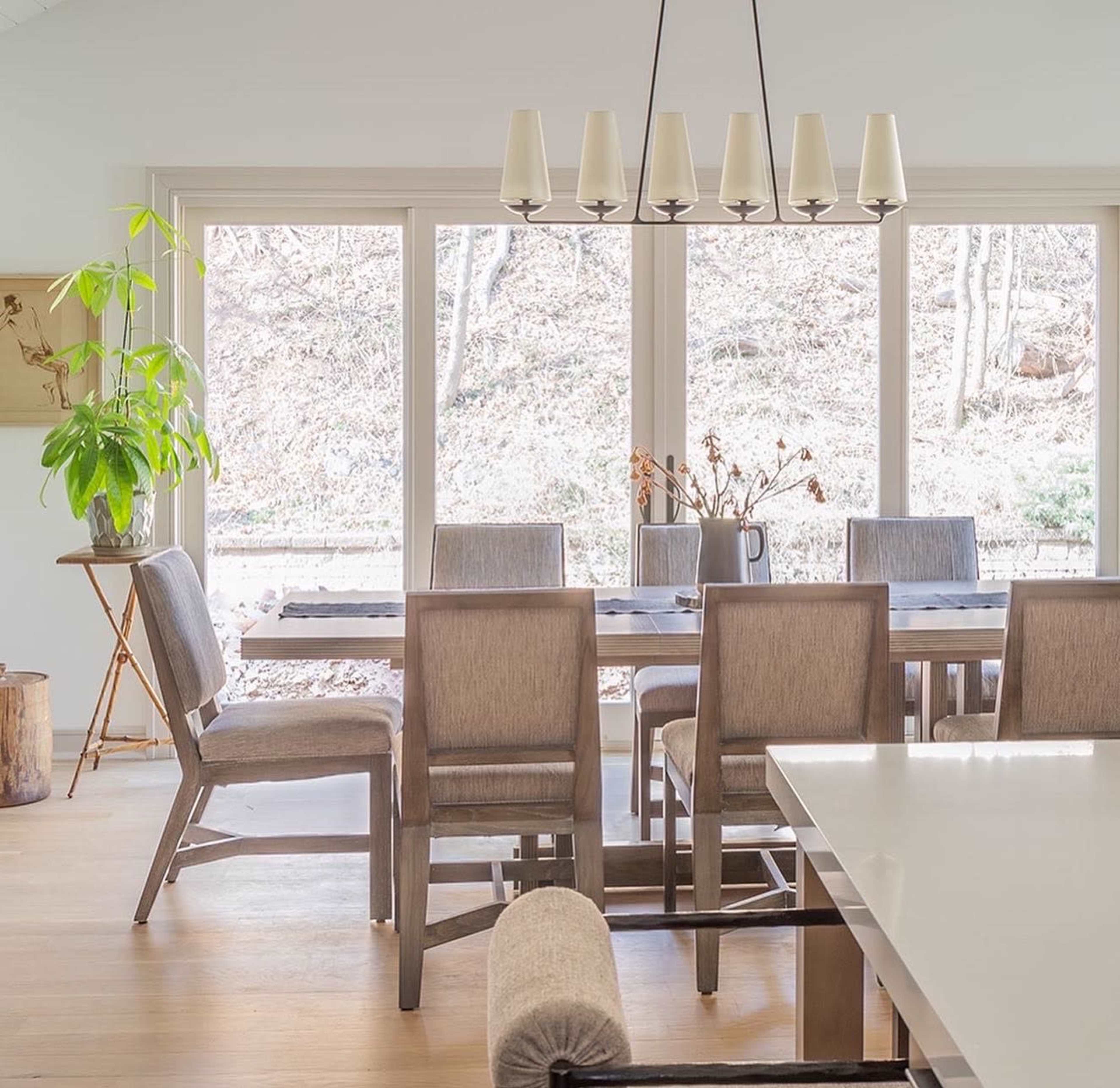 A dining room features a long table surrounded by six chairs, with large windows providing a view of a natural landscape outside.