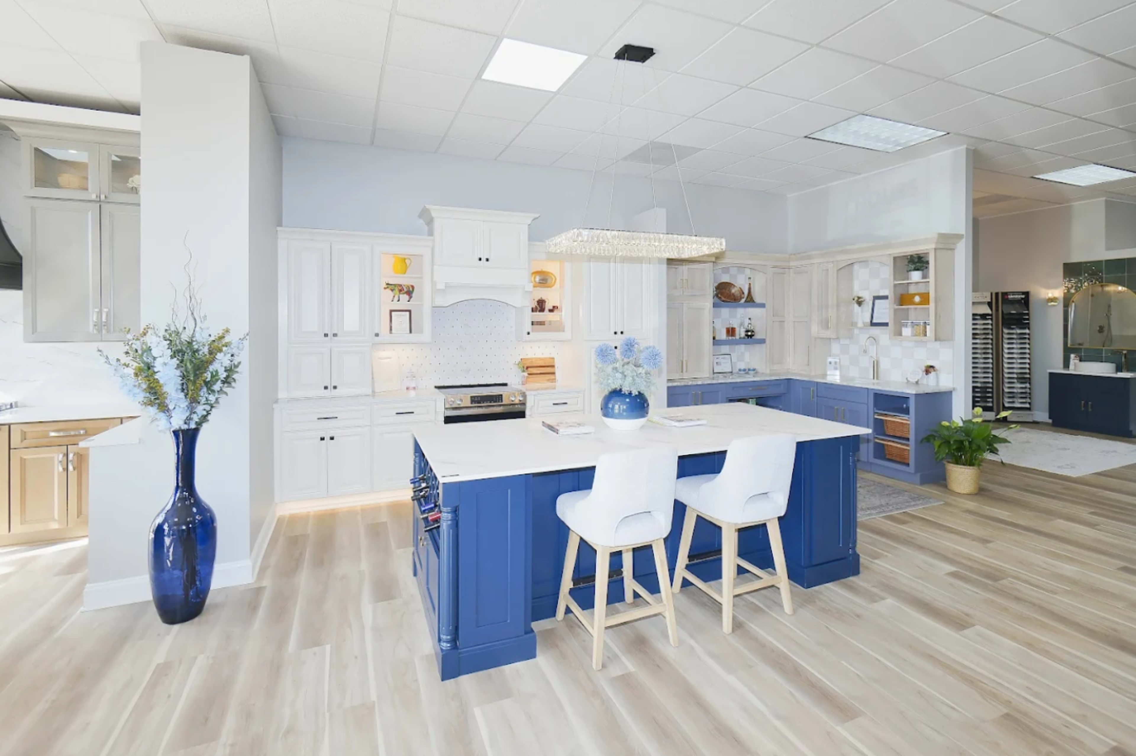 The image shows a modern kitchen showroom featuring white and blue cabinetry, a large island with bar stools, and a decorative vase on the counter.